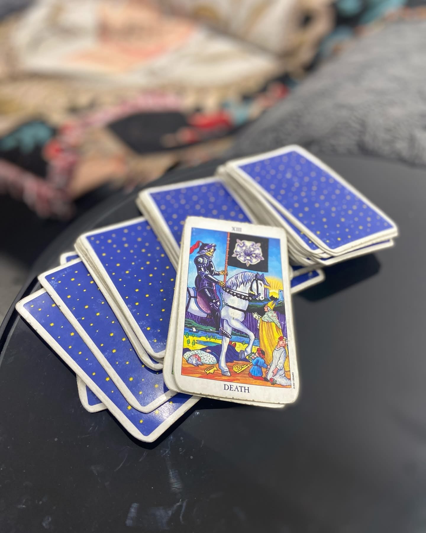 A tarot card titled 'Death' depicting a knight on a white horse, holding a black flag with a white skull, with various characters and scenes around, placed among several other face-down tarot cards with blue backs with gold star patterns.