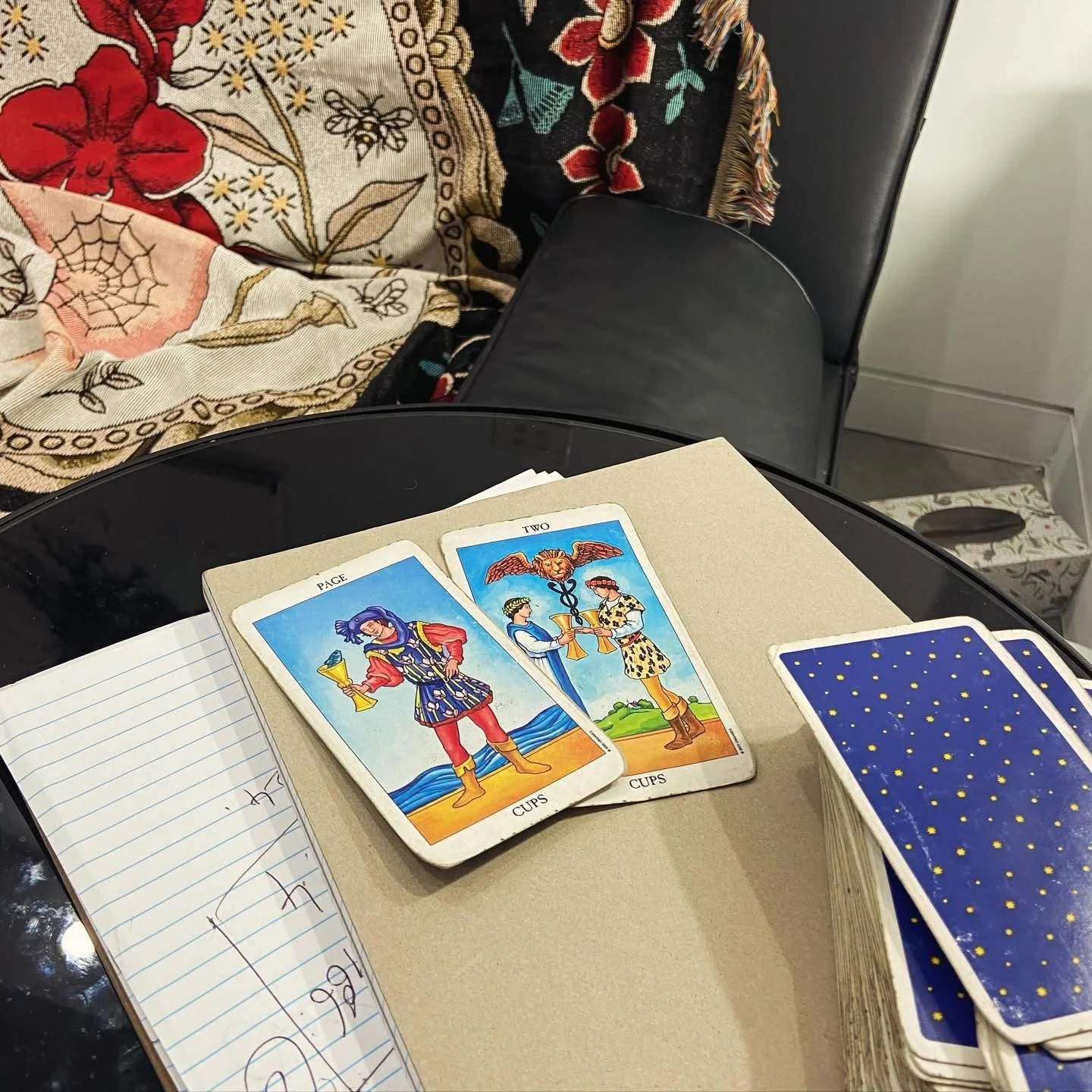 A tarot card reading scene with two tarot cards on a notebook. One card shows a person holding a glass, and the other depicts two people with an eagle and a caduceus. To the right, there is a deck of blue and yellow star-patterned tarot cards. In the background, there's a black chair with a floral and patterned fabric draped over it and a box of tissues on the floor.