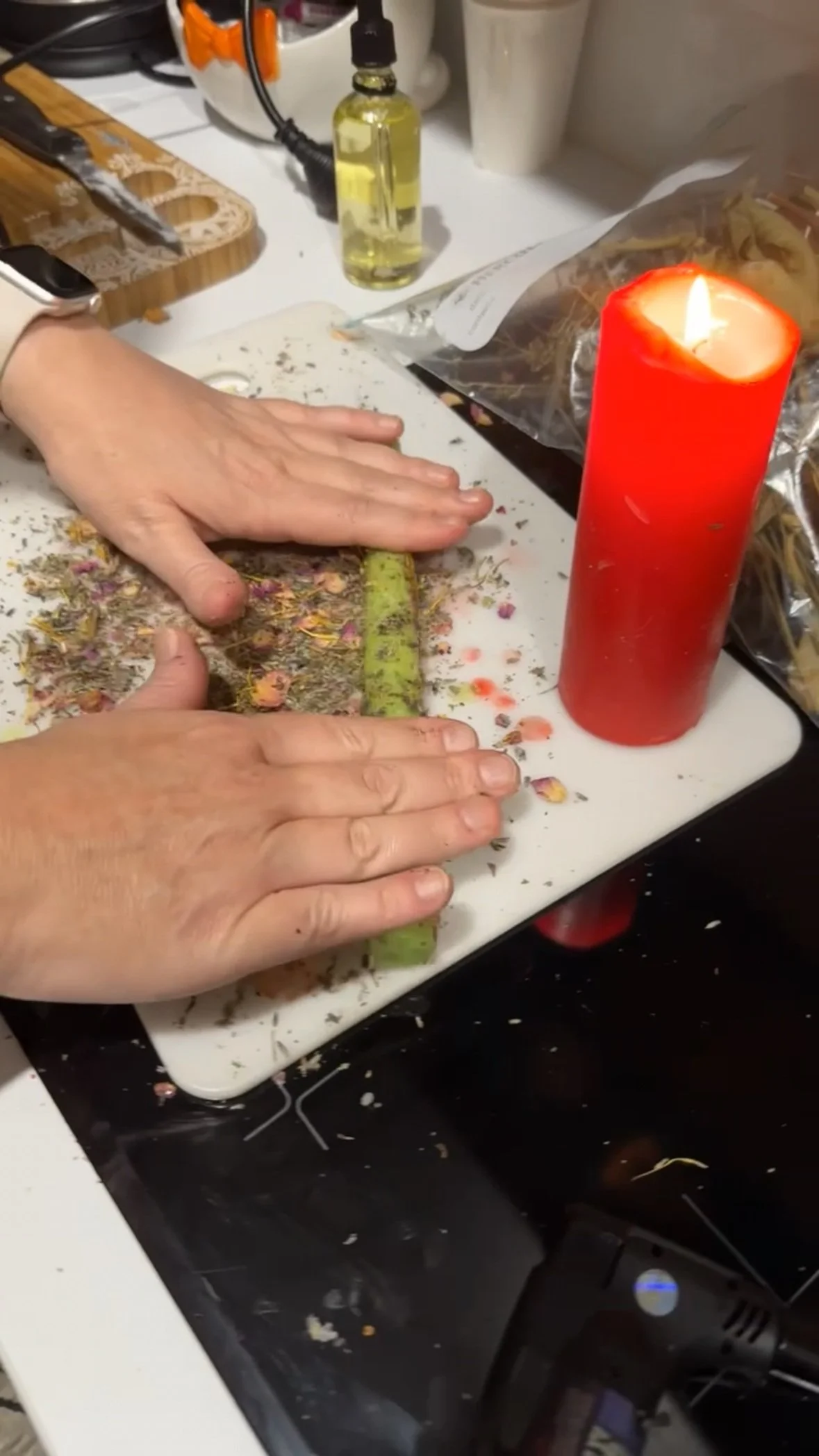 Someone crushing dried flowers and herbs on a white cutting board, with a red candle nearby and various kitchen items in the background.