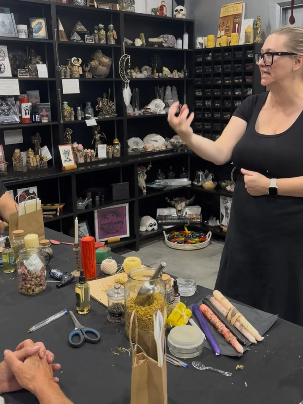 A woman with glasses, wearing a black shirt and apron, gesturing while talking to a person with clasped hands, in a room with shelves filled with crystals, figurines, and decorative objects. The table has craft supplies including candles, yarn, scissors, and bottles.