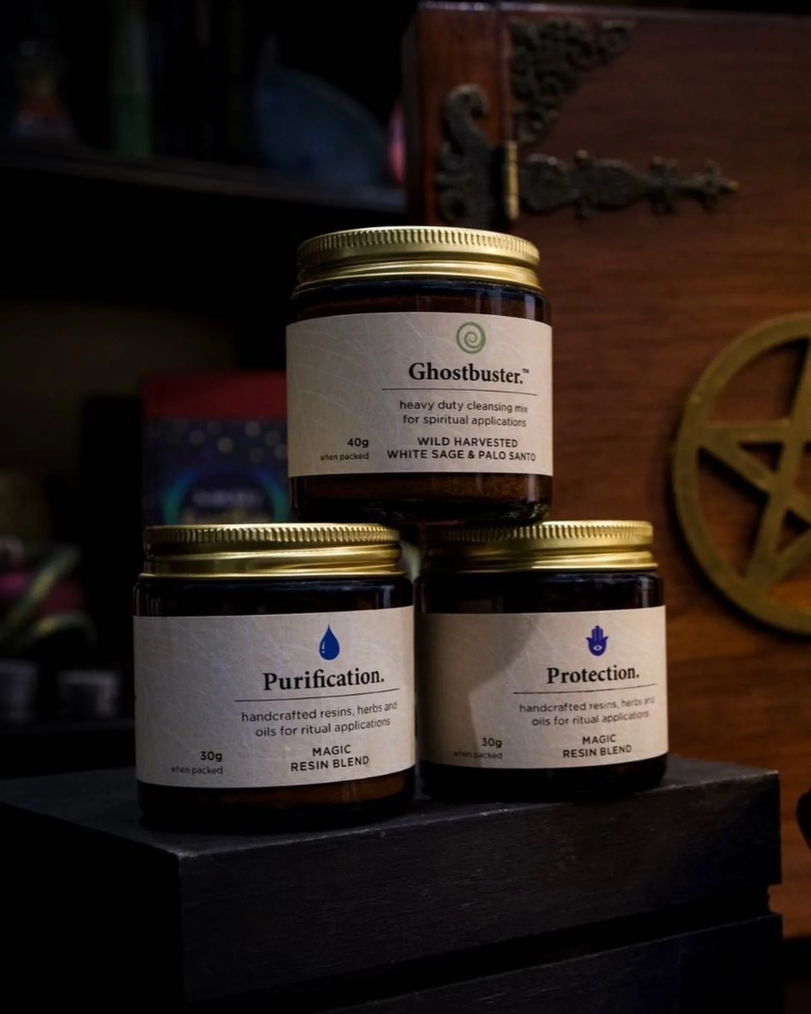 Three jars labeled 'Ghostbusters,' 'Purification,' and 'Protection,' containing resin blends and herbal oils for ritual applications, placed on a dark surface with a wooden background.