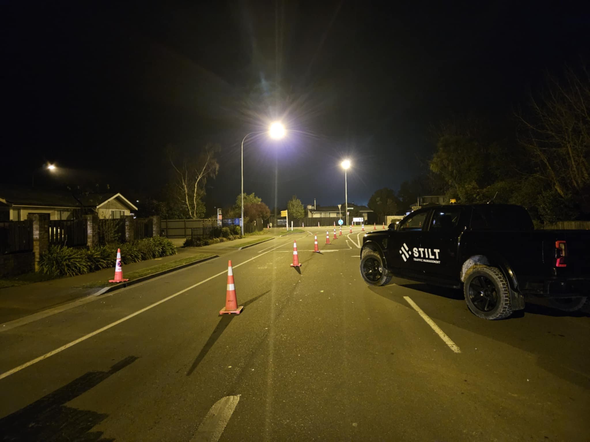 Night scene of an empty street with orange traffic cones set up, a black traffic management vehicle with 'STILT' logo, streetlights illuminating the road, and residential houses on the side.