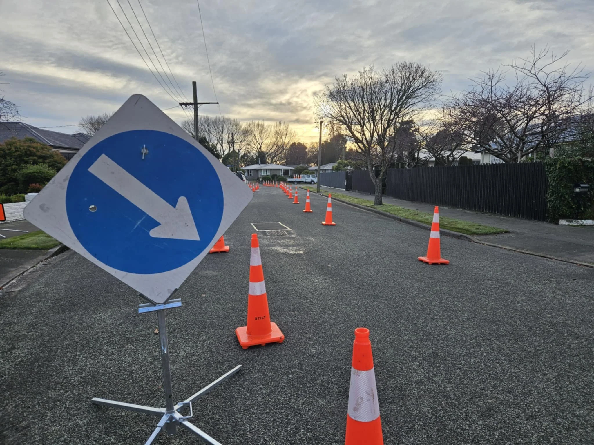 Residential street under construction with orange traffic cones blocking part of the road and a blue circular road sign with a white arrow pointing to the left.