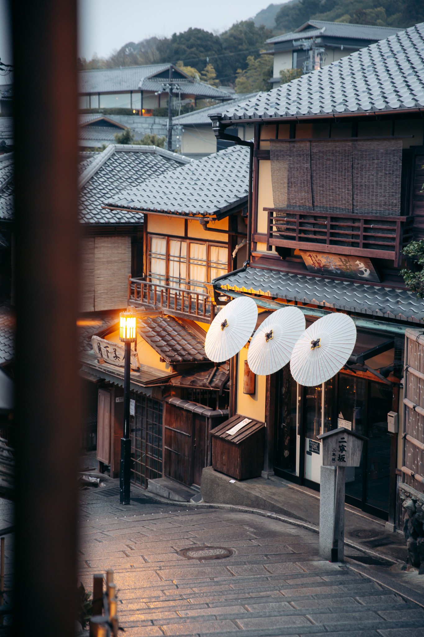 Traditional Japanese street scene with wooden buildings, tiled roofs, paper umbrellas, and a streetlamp, during evening.