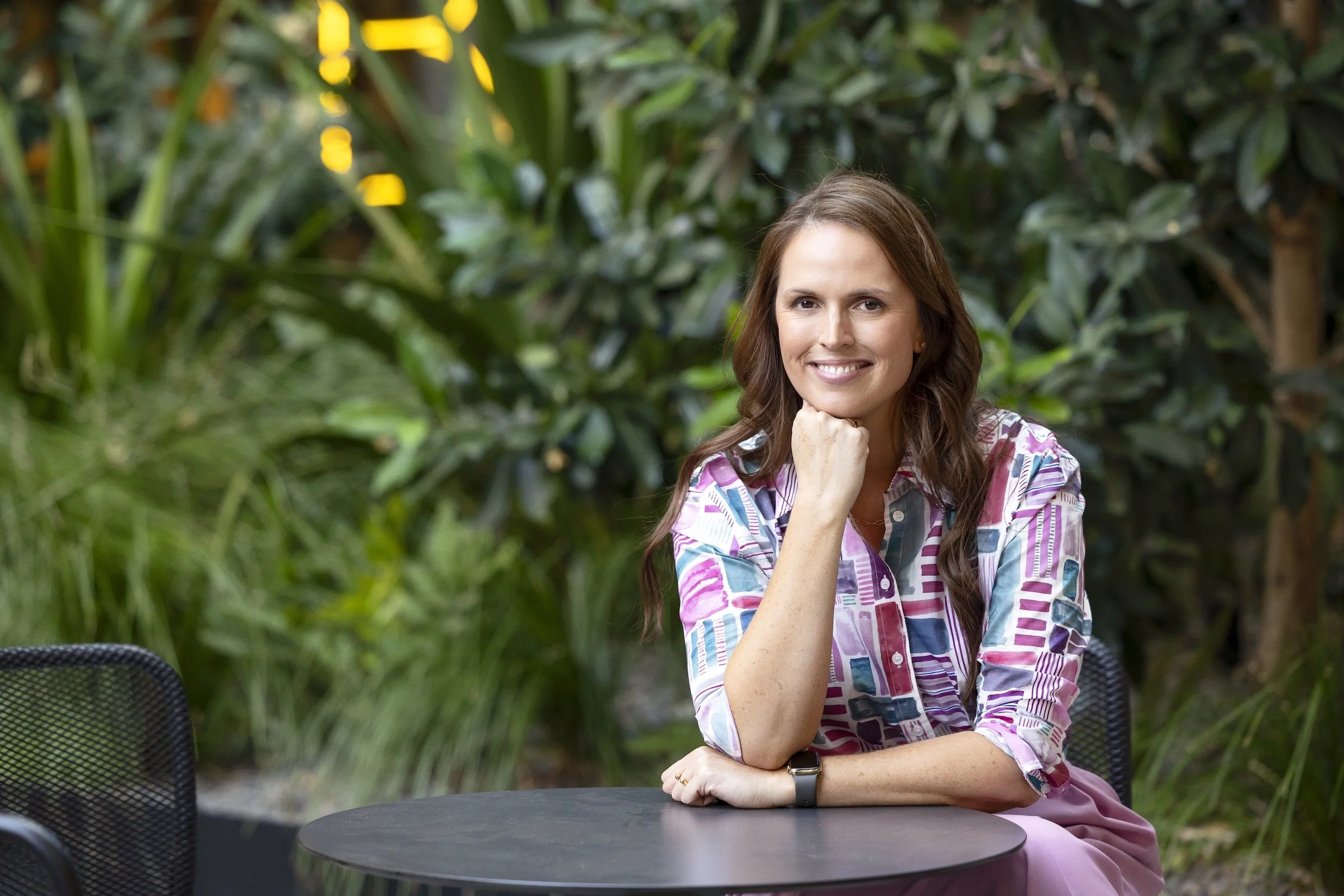 Photo of Laura Kendall, seated at an outdoor table in front of a background of greenery. Laura smiles at the camera. She is a white, middle-aged woman with brown eyes and long brown hair, wearing a shirt with a pink, white, green geometric pattern.