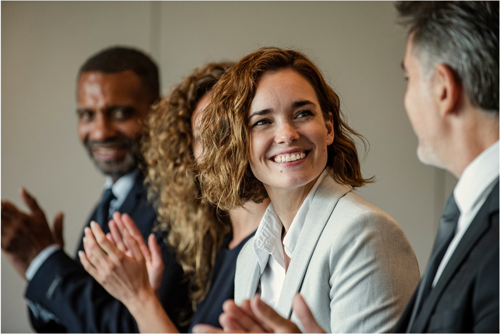 A woman with curly red hair, wearing a beige blazer and white shirt, is smiling and engaged in conversation with a man. Other people are clapping in the background during what seems to be a professional event or meeting.