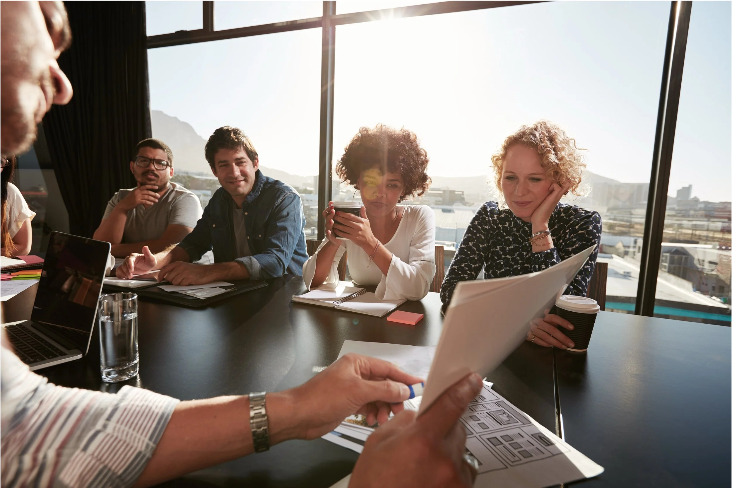 Photo of several colleagues seated around a table in a sunlit boardroom. One is pointing to a page, while others watch