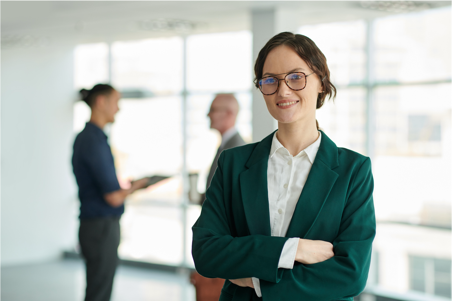 Photo of a white woman in professional attire smiling at camera with a blurred background where two colleagues stand facing each other in conversation