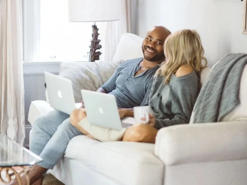 A man and a woman sitting on a sofa in a living room, smiling and talking. The woman is holding a mug and a laptop, the man has a laptop on his lap.