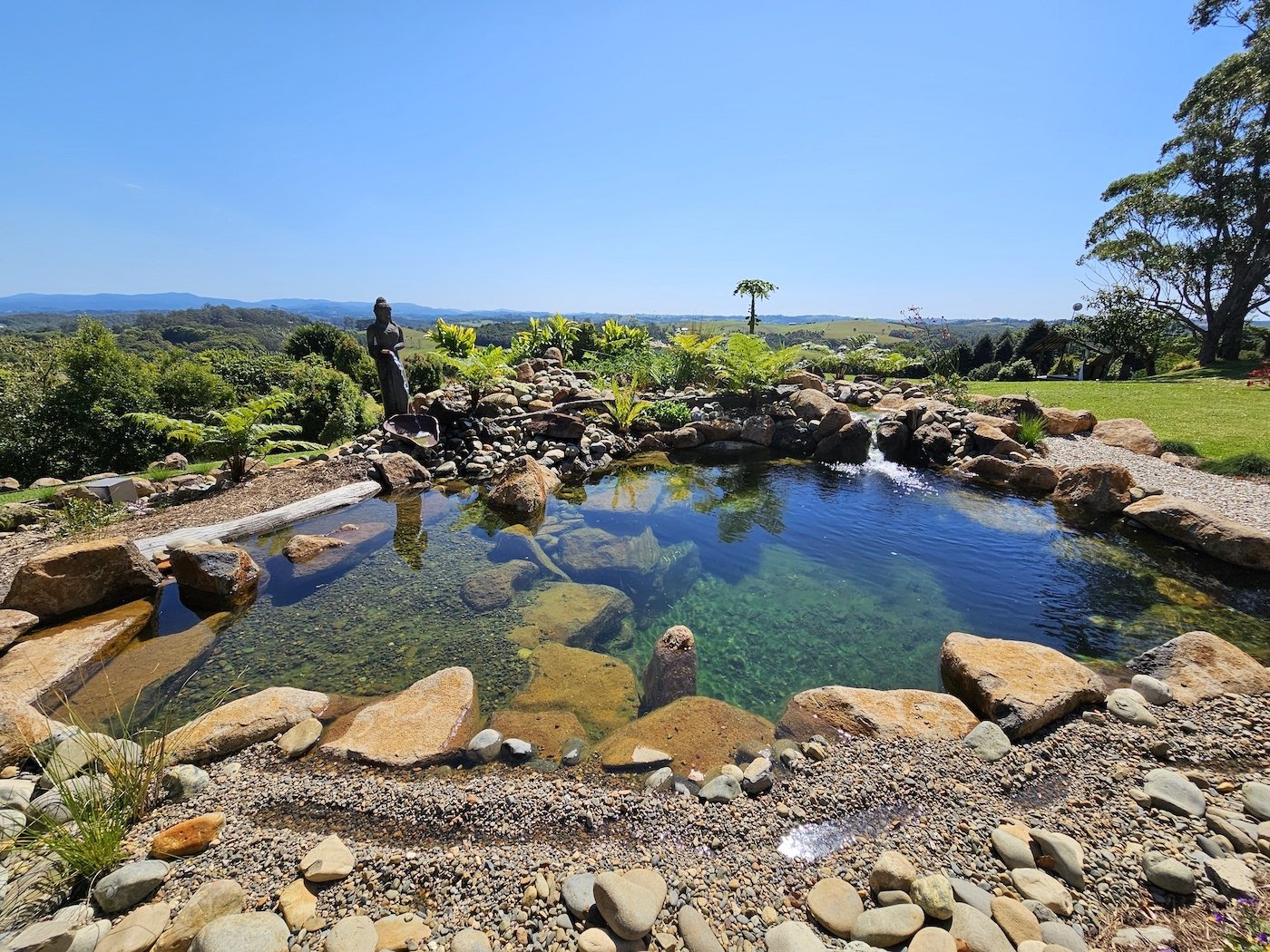 Panoramic view of a flo garden natural swimming pool in Byron Bay surrounded by rocks, a few plants, and the statue of a middle size Buddha