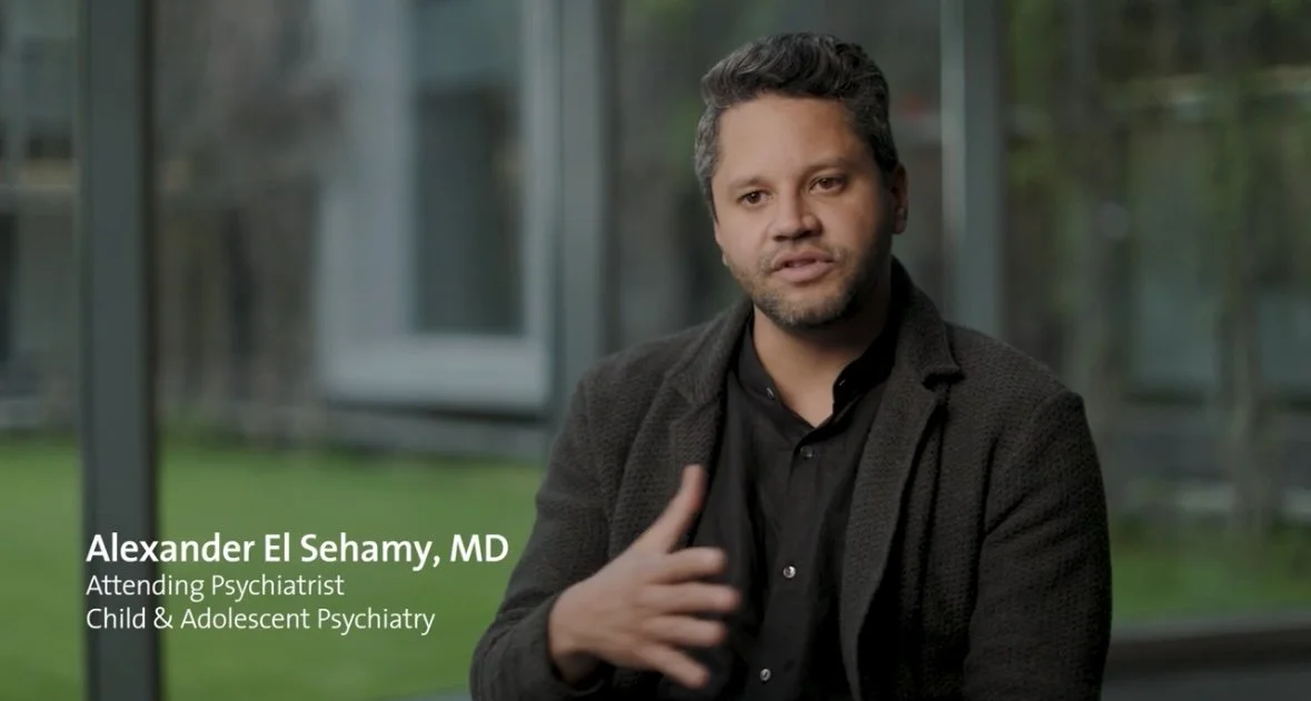 A man with dark and gray hair, wearing a black blazer and shirt, sitting indoors near a window with a blurred green outdoor background, speaking to the camera. Text overlay indicates his name is Alexander El Sehamy, MD, an attending psychiatrist in child and adolescent psychiatry.