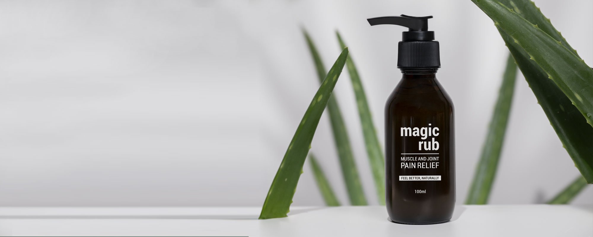 A brown bottle labeled 'magic rub' with a pump dispenser, placed on a white surface with green aloe vera leaves in the background.