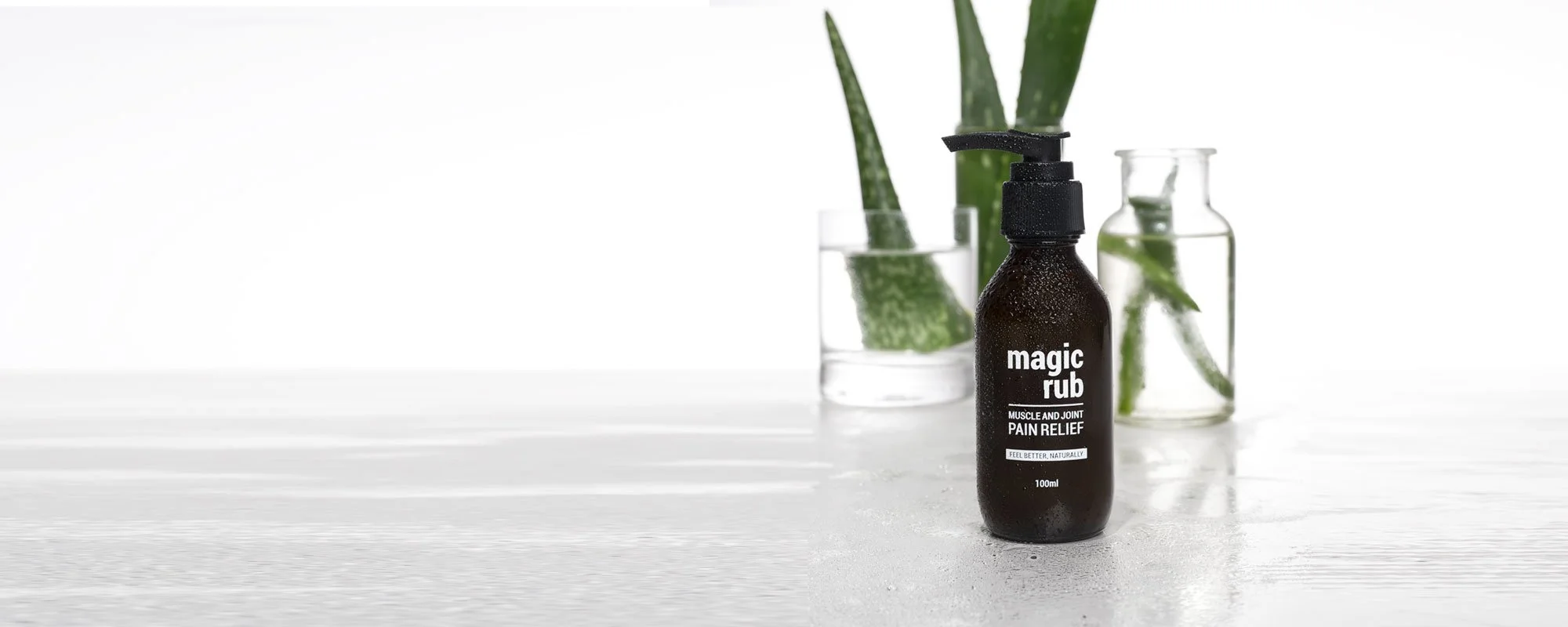 A spray bottle labeled 'magic rub' for muscle and joint pain relief placed on a white surface, with glass containers filled with aloe vera and water in the background.