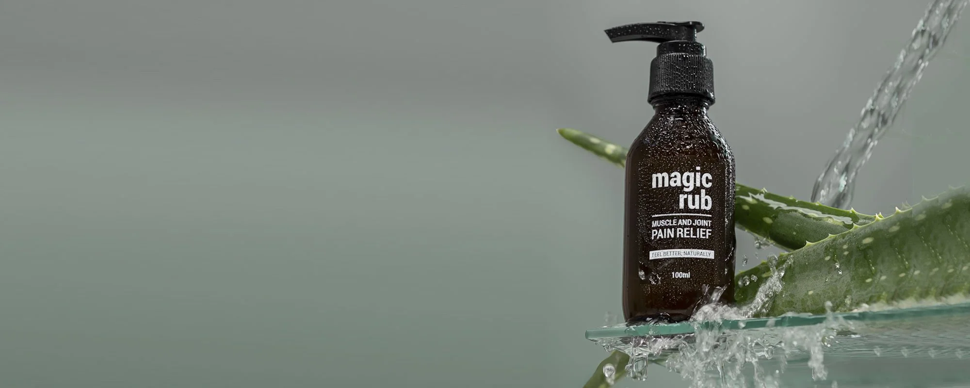A brown spray bottle labeled 'magic rub muscle and joint pain relief' with a pump top, placed on a reflective surface with aloe vera leaves and water splashing around.