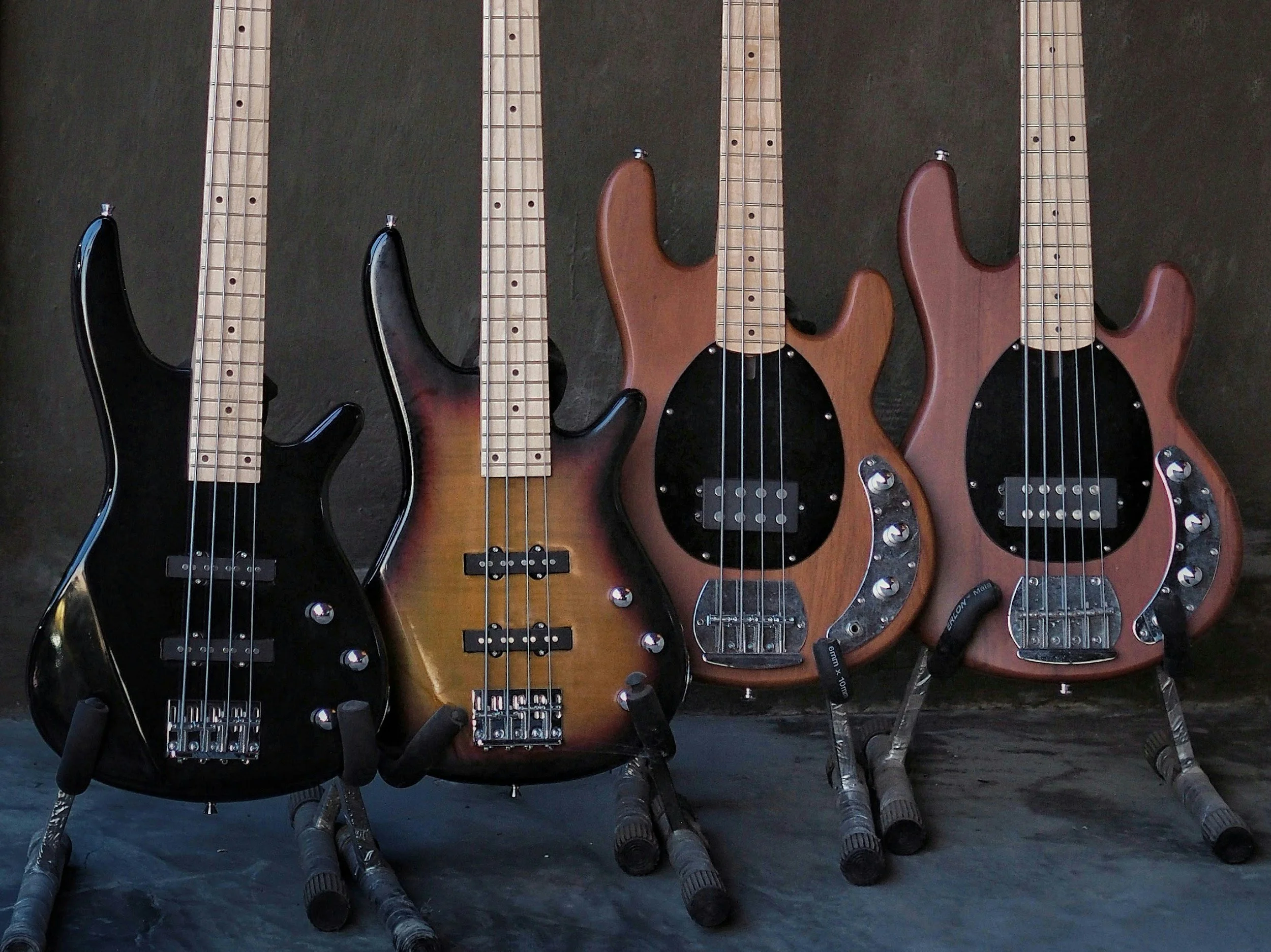 Four electric bass guitars, two black, one sunburst, and one natural wood finish, arranged on stands against a dark background.