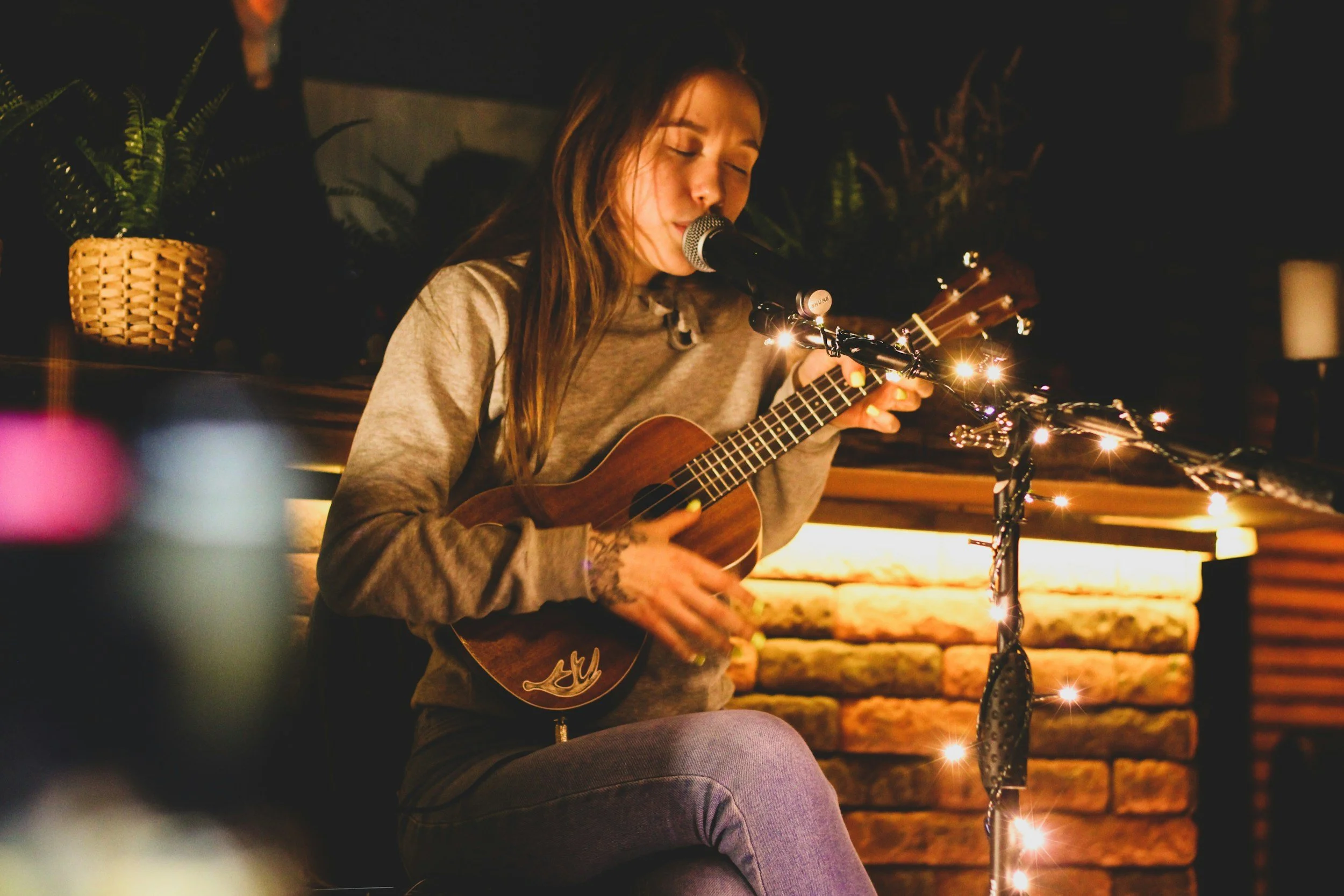 Young woman with long hair, eyes closed, singing into a microphone while playing an acoustic guitar on a dimly lit stage decorated with string lights and plants.