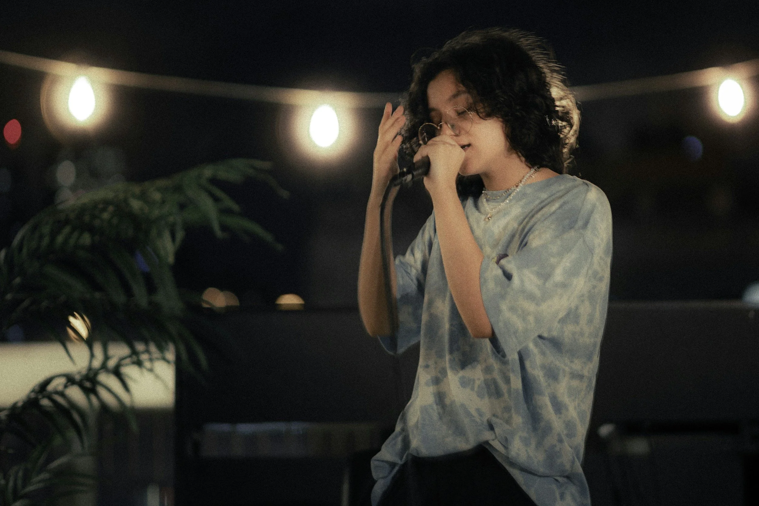 Young woman with curly hair and glasses performing with microphone at night, illuminated by string lights, with a plant in the foreground.