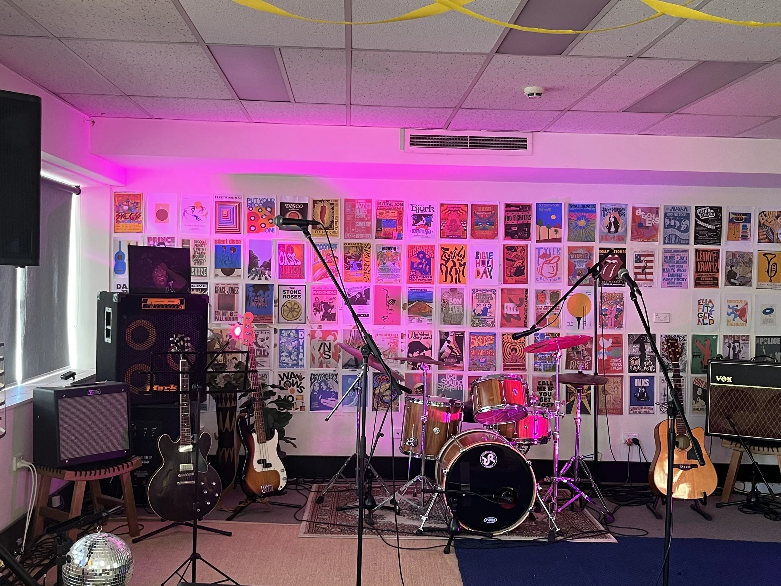 Music studio setup with a keyboard on a stand, an amplifier, a microphone, headphones, a mixing console, and some potted plants near large windows, with colorful abstract artwork on the wall.