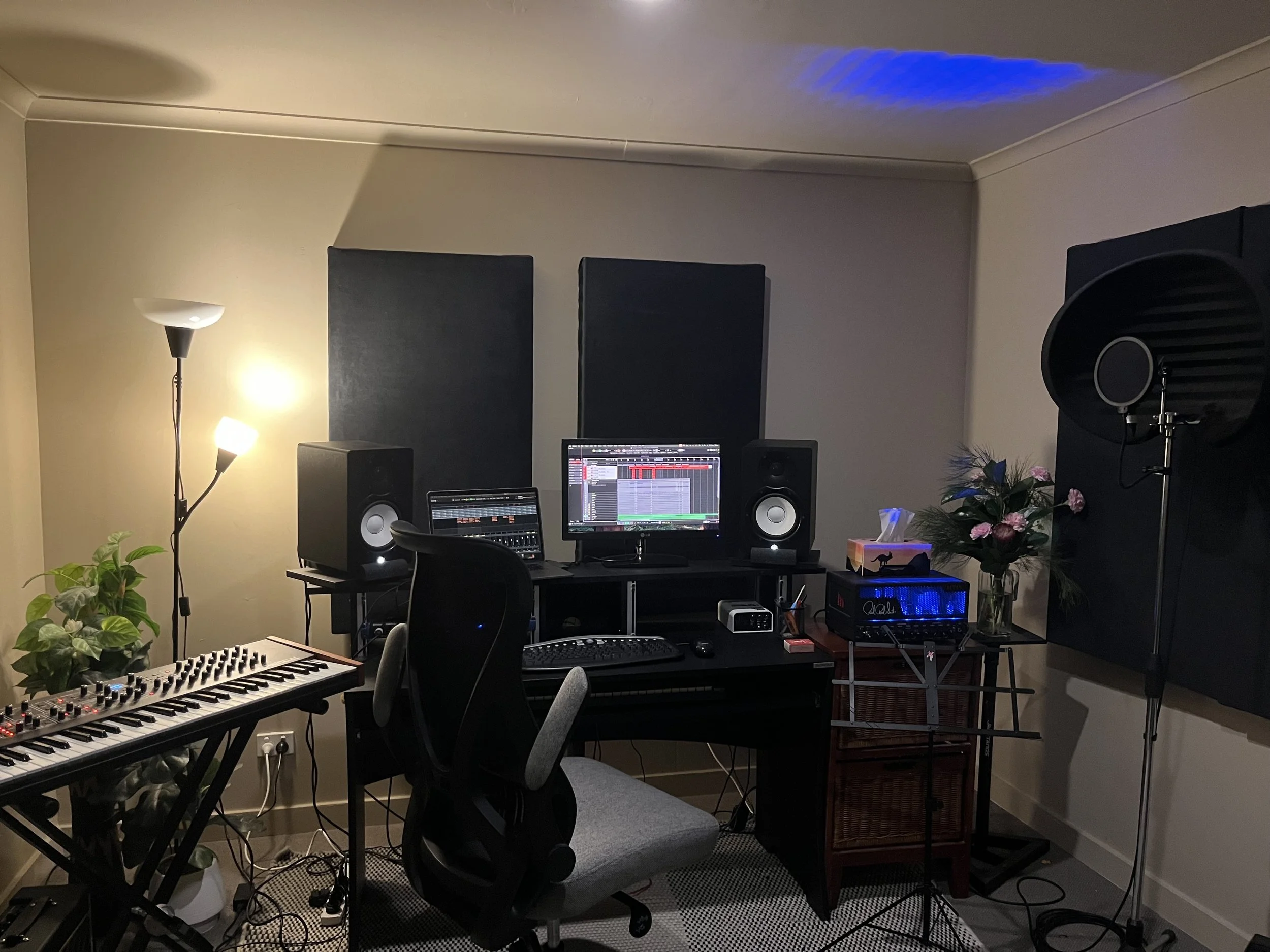 Music recording studio with drums, keyboard, guitar, speakers, mixing equipment, chair, and decor.