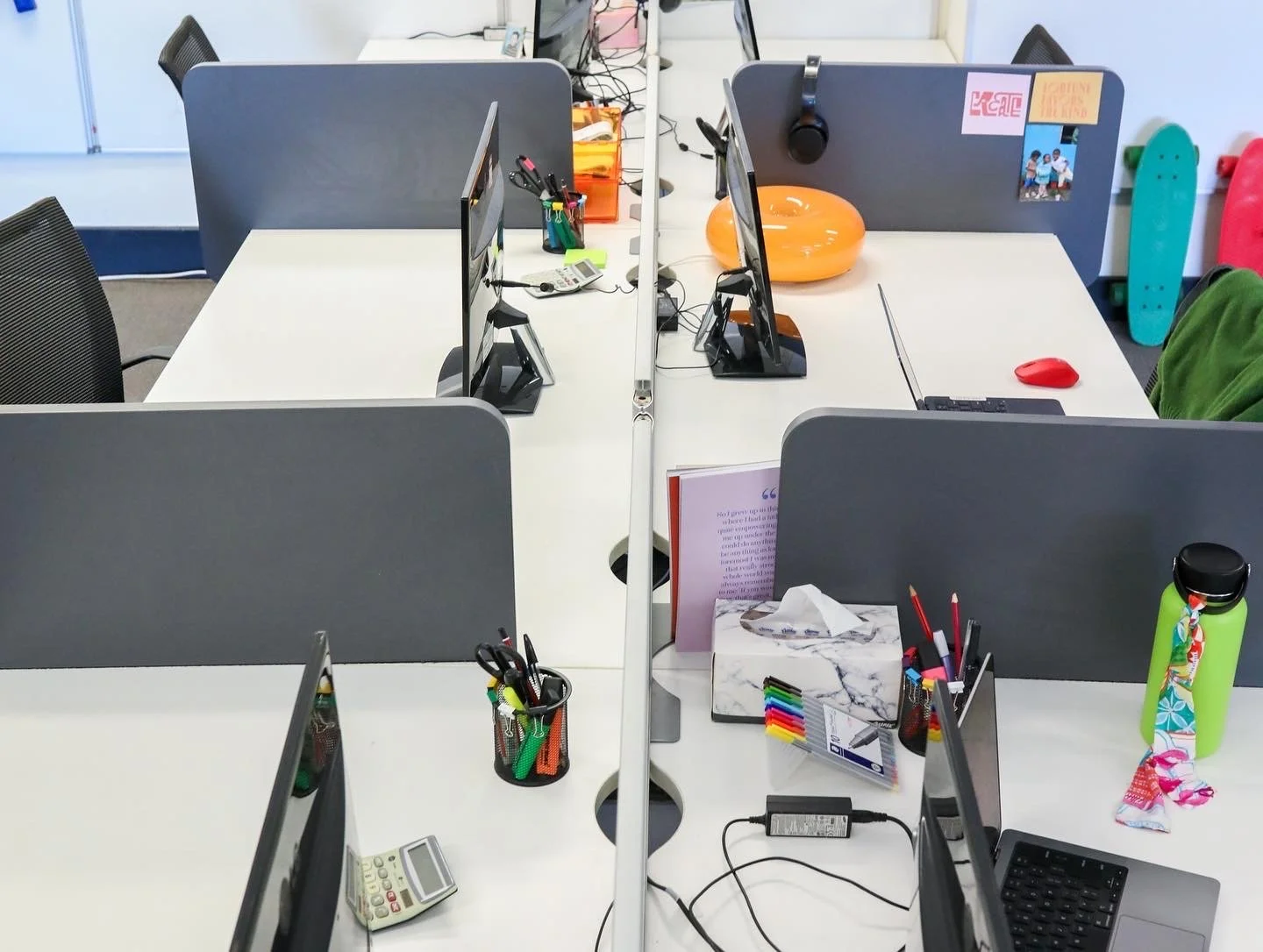 Empty office cubicles with multiple computer monitors, chairs, and some office supplies, with a white desk and partition screens.