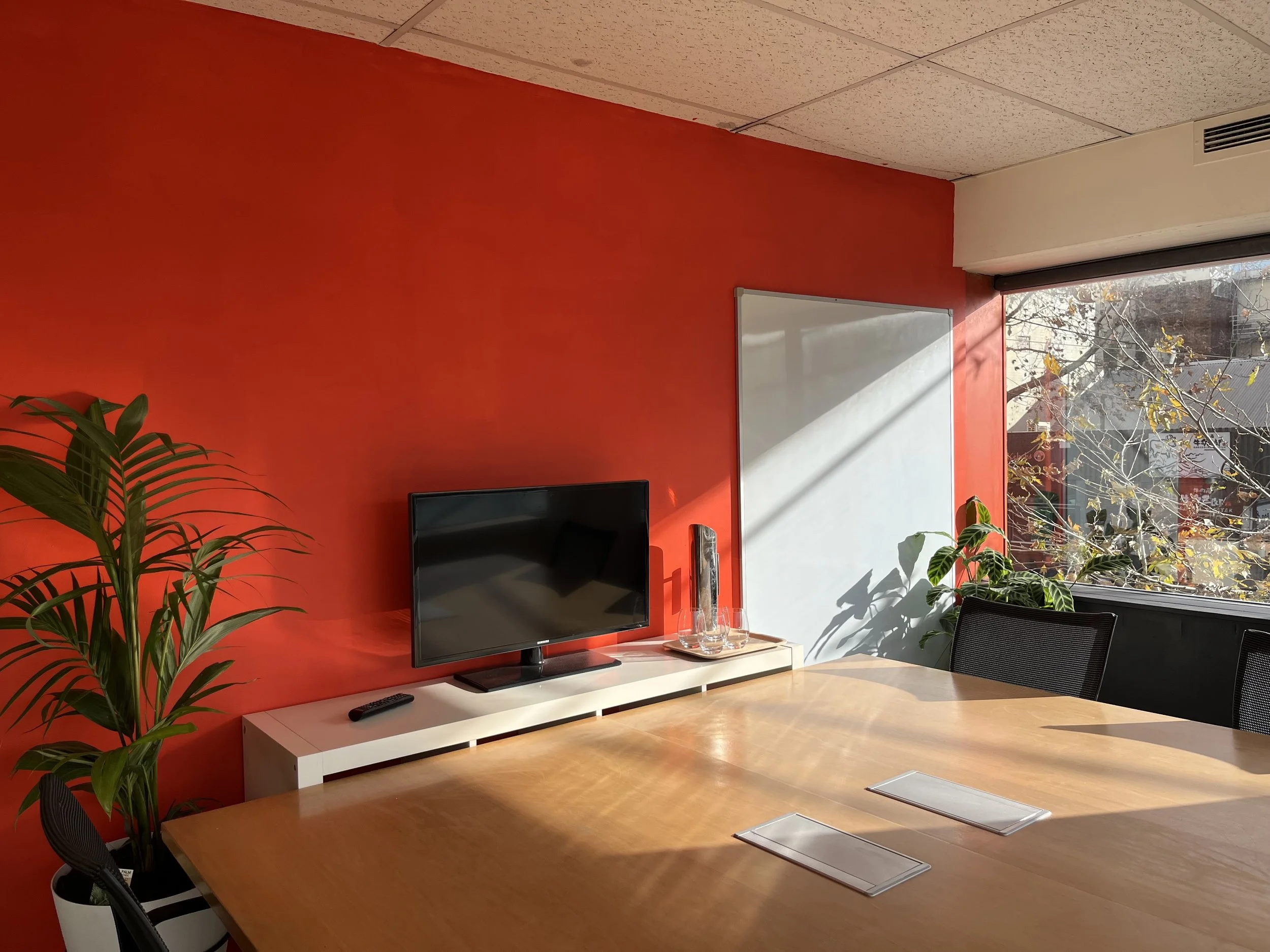 Empty conference room with a wooden table, black chairs, a large television, whiteboard with markers, and a window showing trees outside.