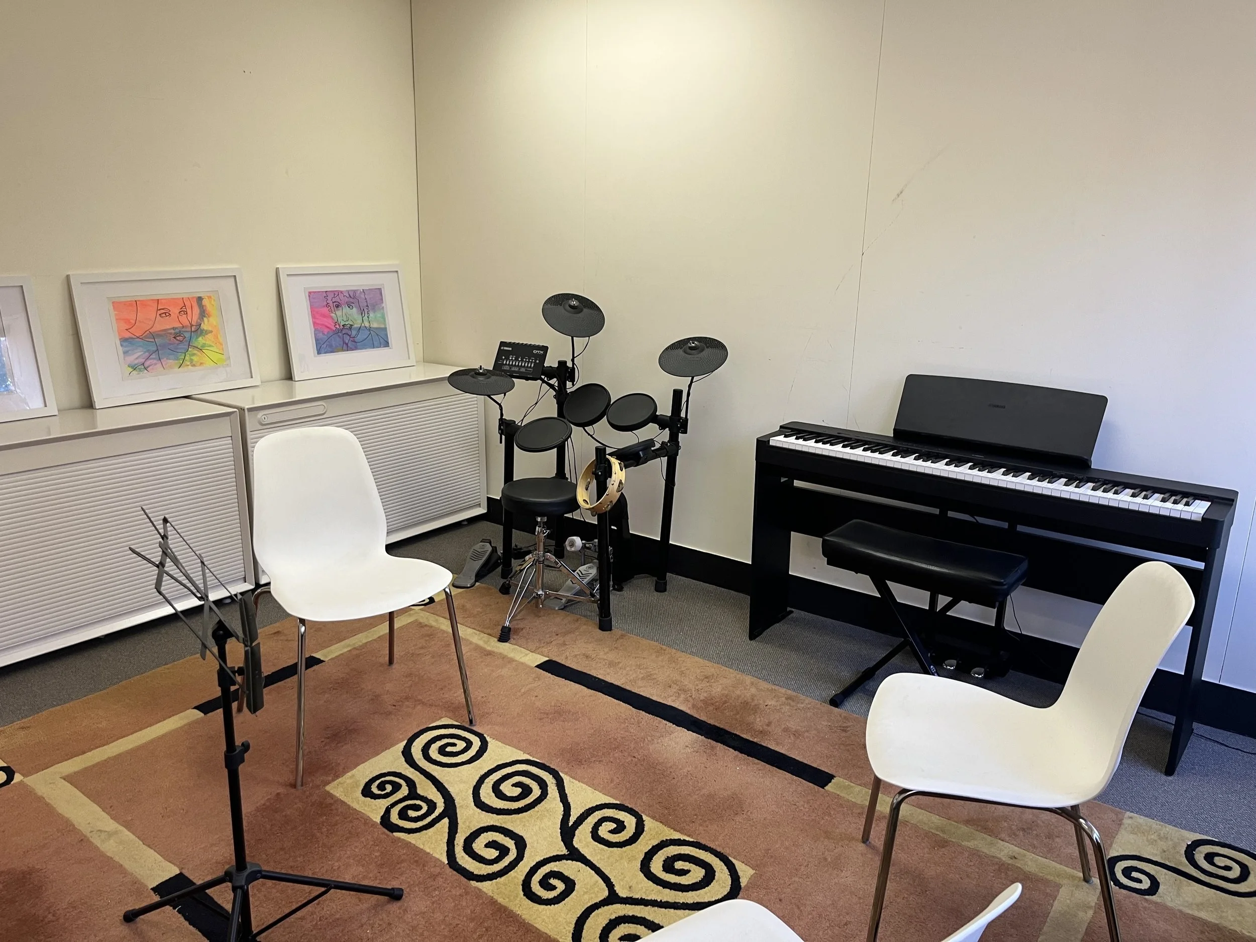 Room with musical instruments including a digital piano, drum set, and amplifiers, with chairs and music stands, a rug, and a window with trees outside.