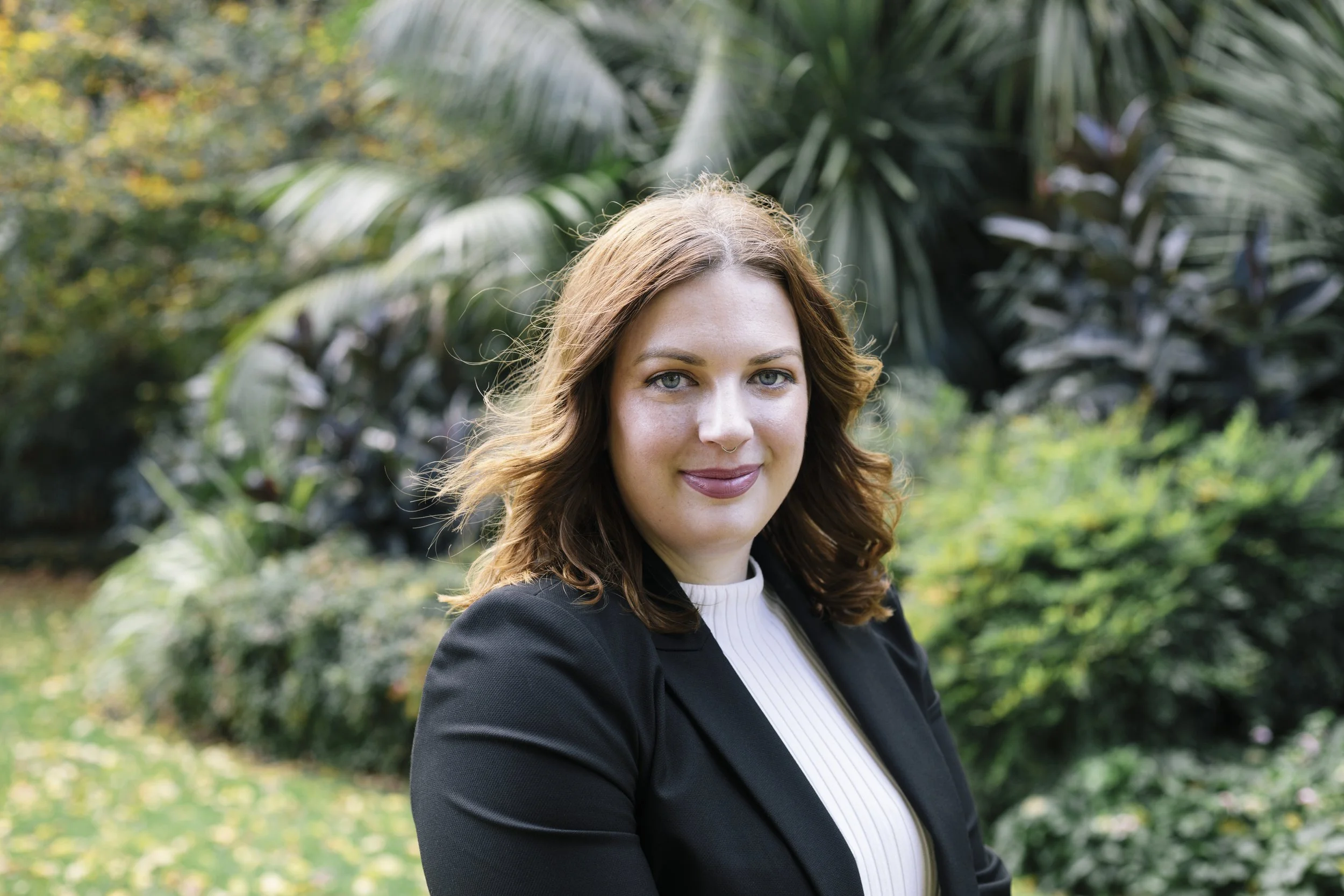 A woman with shoulder-length wavy reddish-brown hair, wearing a black blazer and a light-colored turtleneck, standing outdoors in a garden with lush green plants and trees.