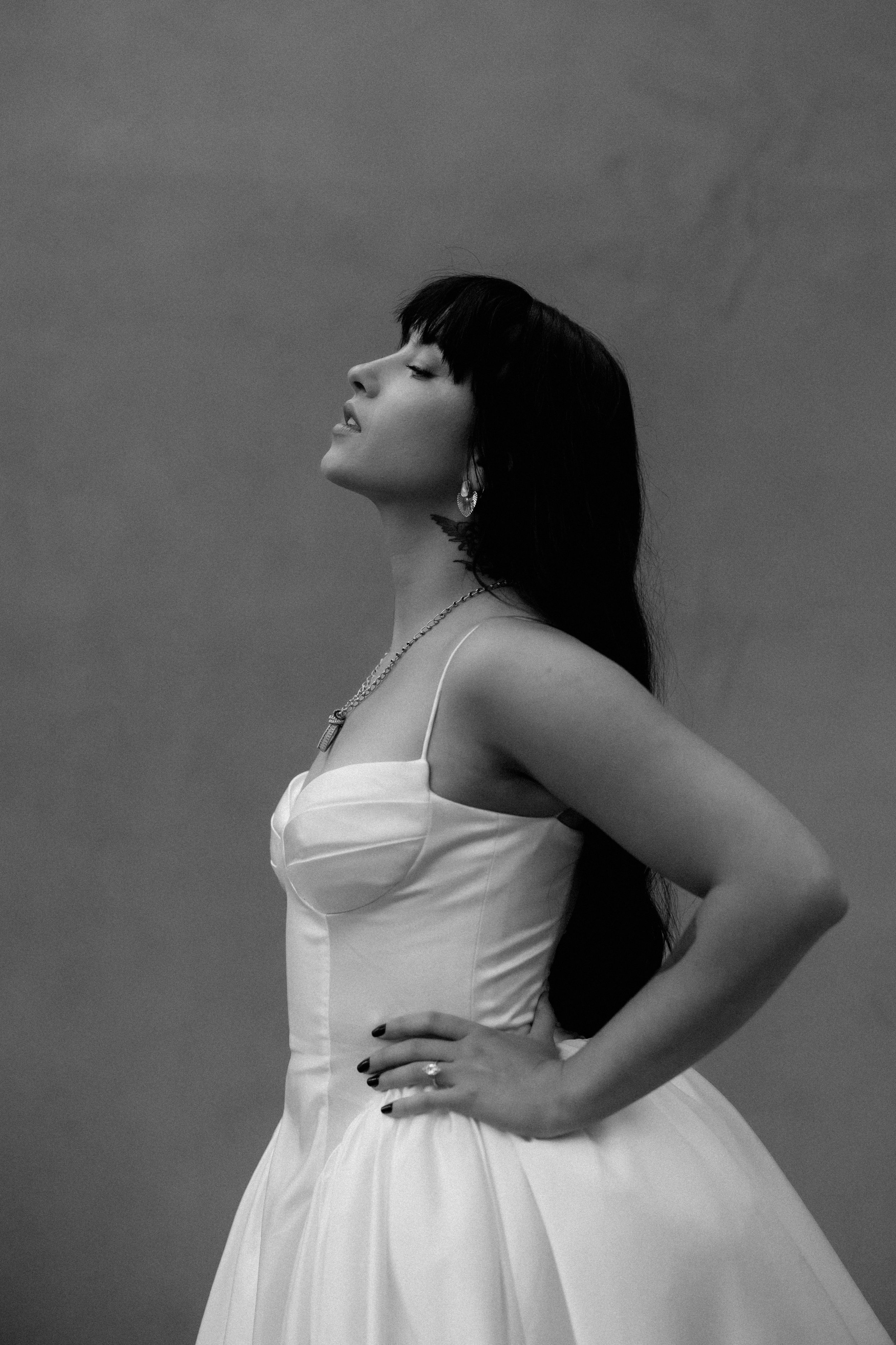 A black and white photo of a woman with long dark hair in a strapless dress, posing with her hand on her hip against a plain background.