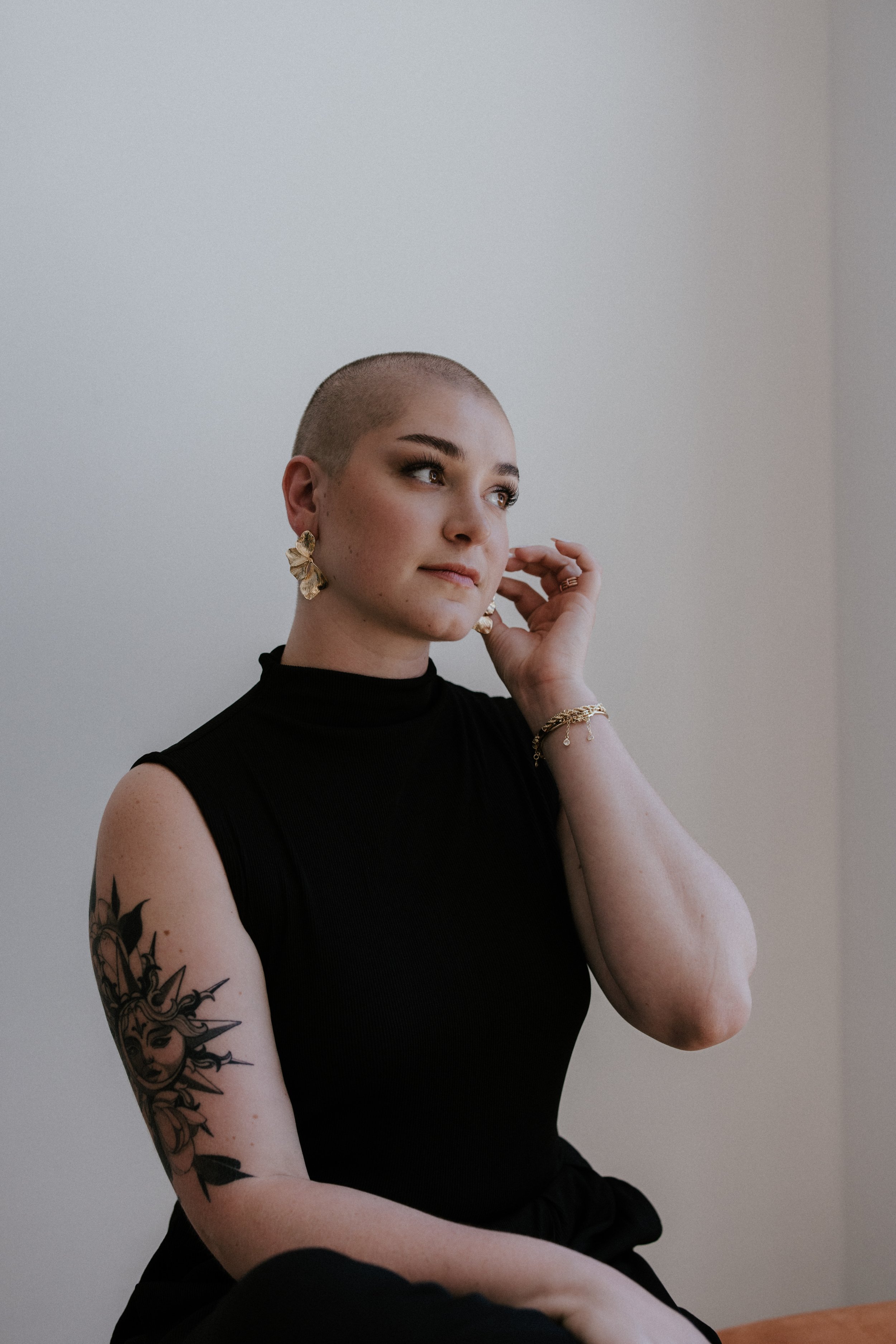 A young woman with a shaved head wearing a sleeveless black top, gold earrings, bracelet, and a tattoo on her left arm, sitting near a plain wall, looking thoughtfully to the side.