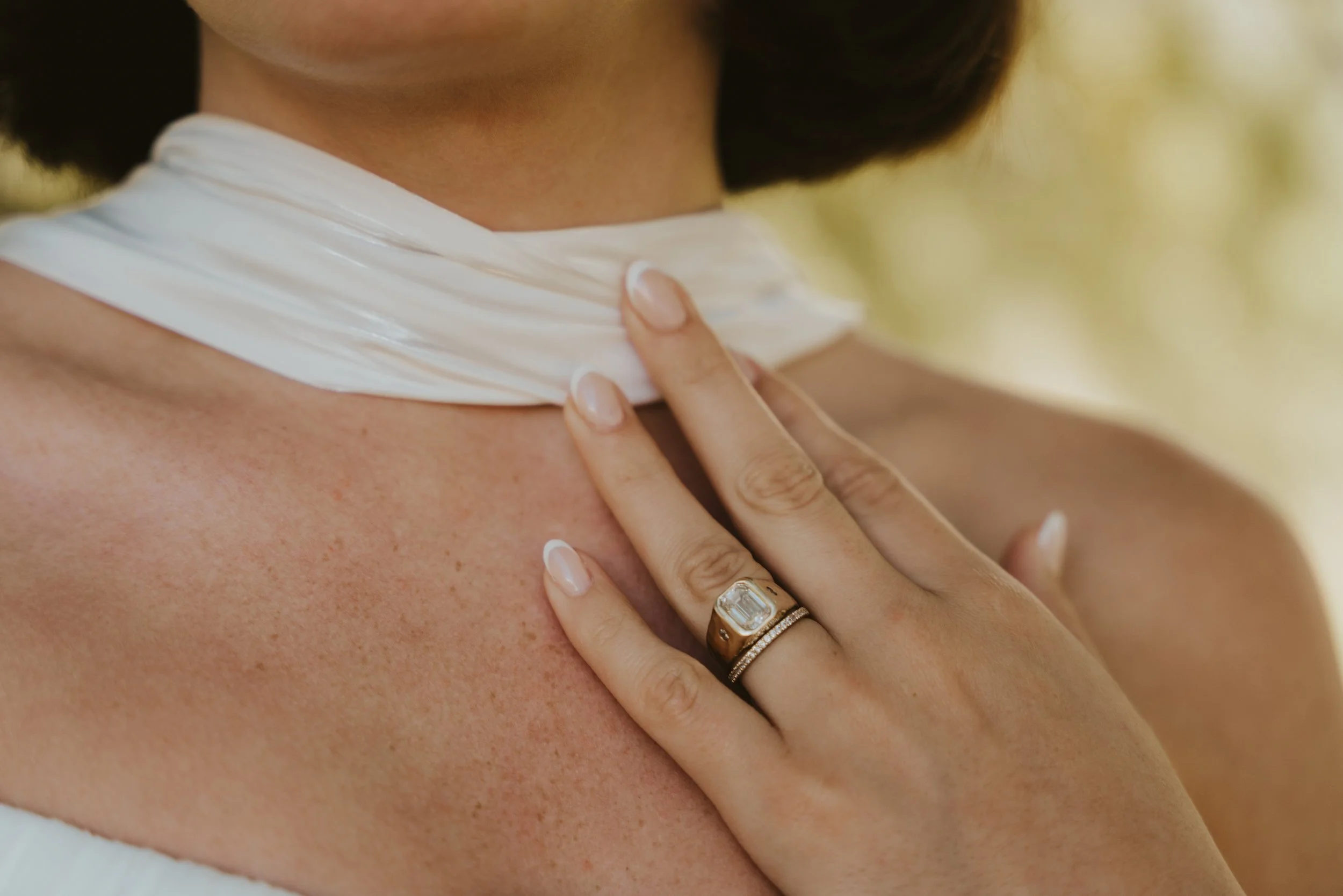 Close-up of a woman's neck and shoulder with her hand resting on her neck. She is wearing a large engagement ring with a prominent rectangular diamond and a wedding band with smaller diamonds. She has a smooth, well-manicured hand with a French manicure. The background is blurred.