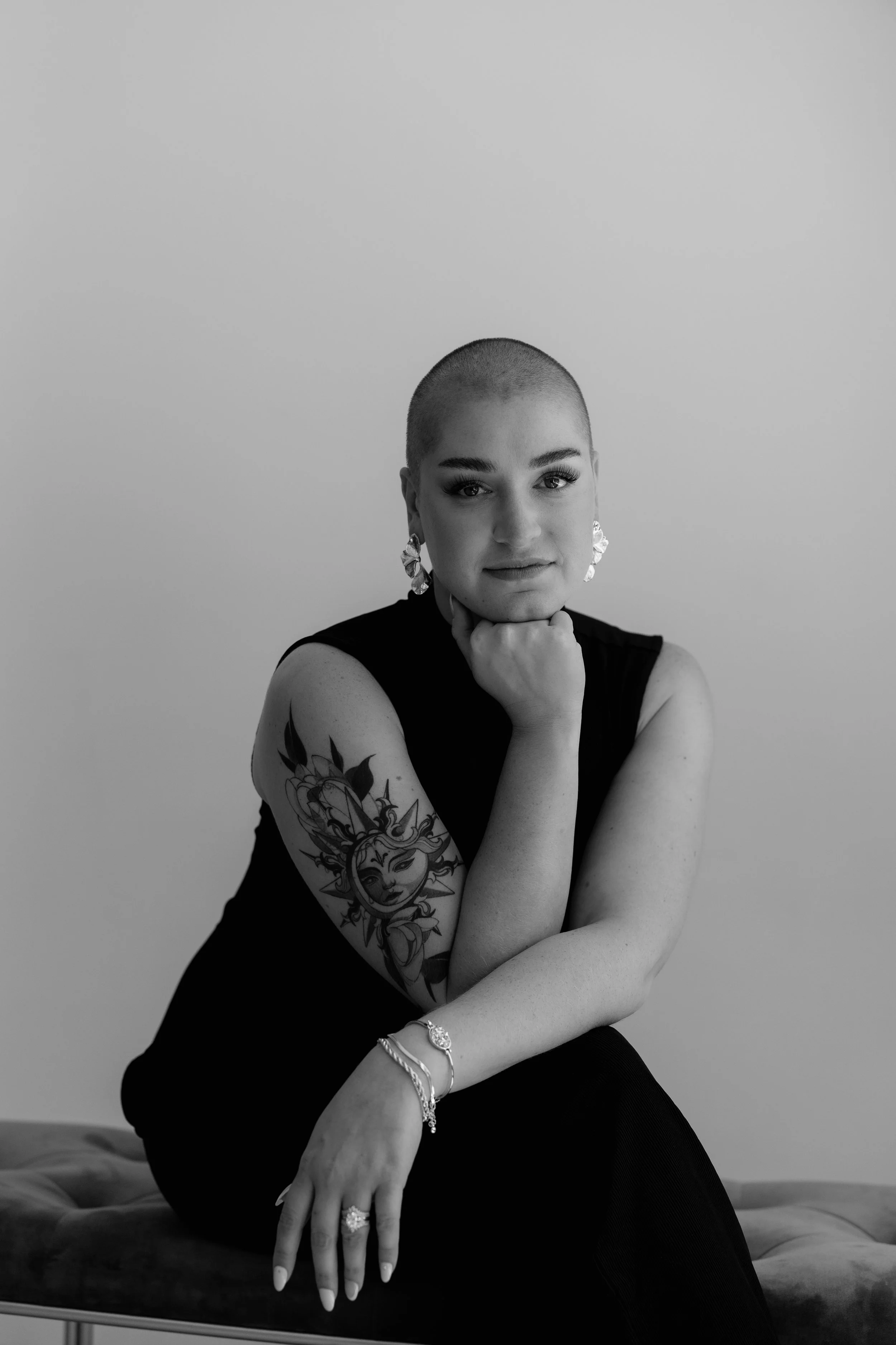 A black-and-white portrait of a woman with a shaved head, wearing earrings, a sleeveless top, and jewelry, sitting with her chin resting on her hand, showcasing tattoos and jewelry.