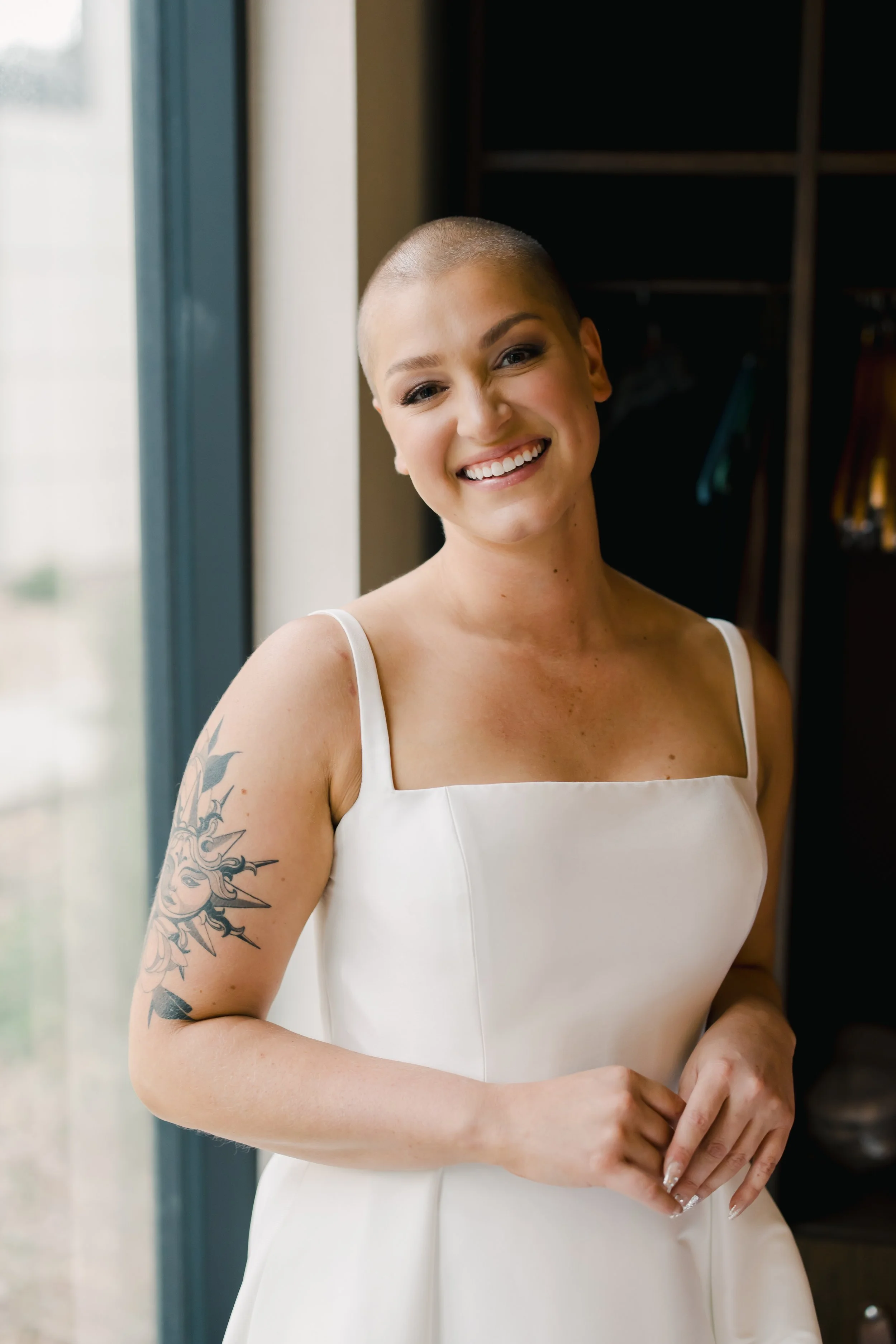A woman with a shaved head wearing a white dress, smiling and standing near a window with daylight shining through.