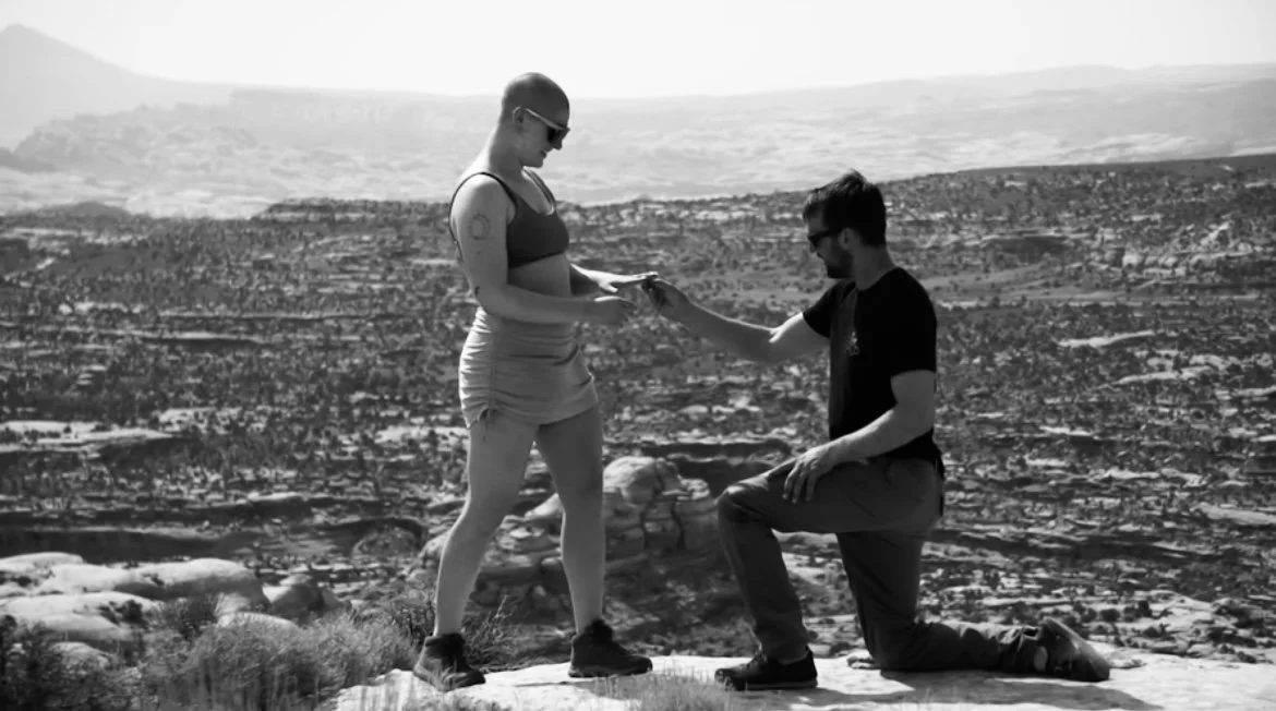 A man proposing marriage to a woman on a scenic overlook with a cityscape background.