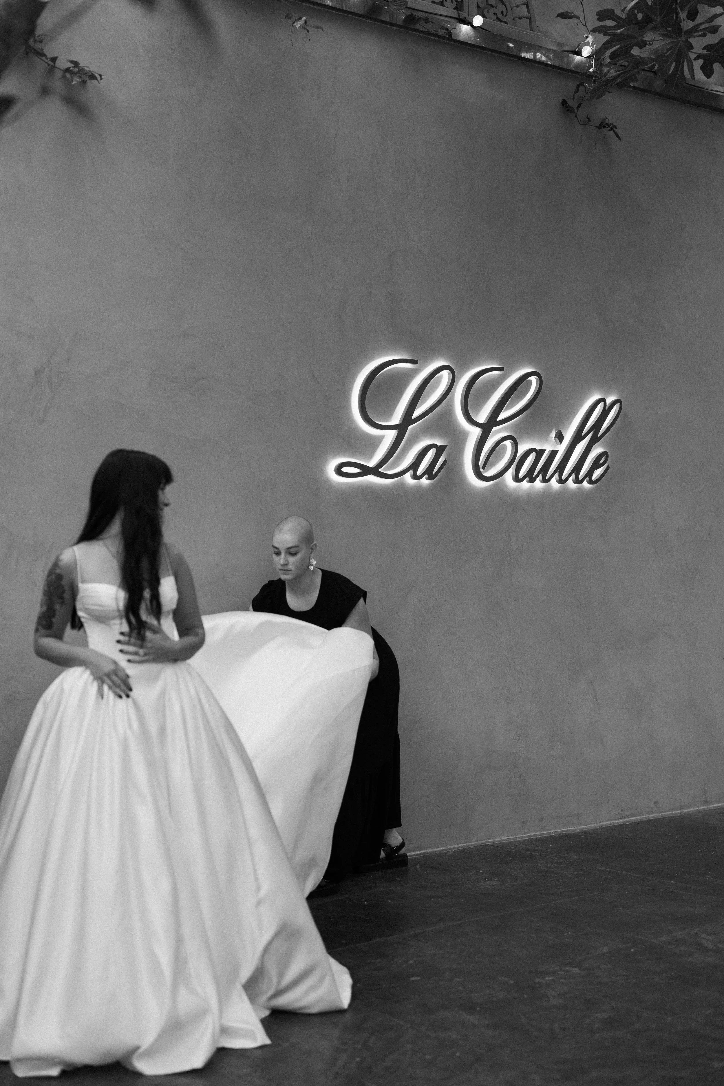 Two women, one in a wedding dress and the other in black, standing near a wall with a illuminated sign that reads "La Calle."