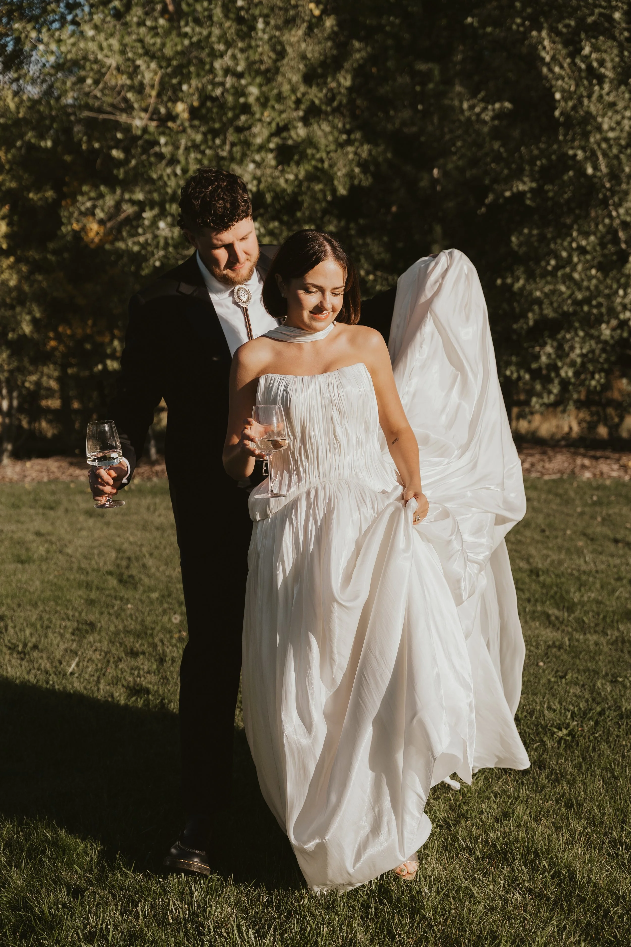 A bride and groom walking outdoors on a grassy area, holding glasses of wine. The bride is smiling, wearing a white dress, and holding up part of her gown. The groom is dressed in a dark suit with a bolo tie, and they are enjoying a moment together.
