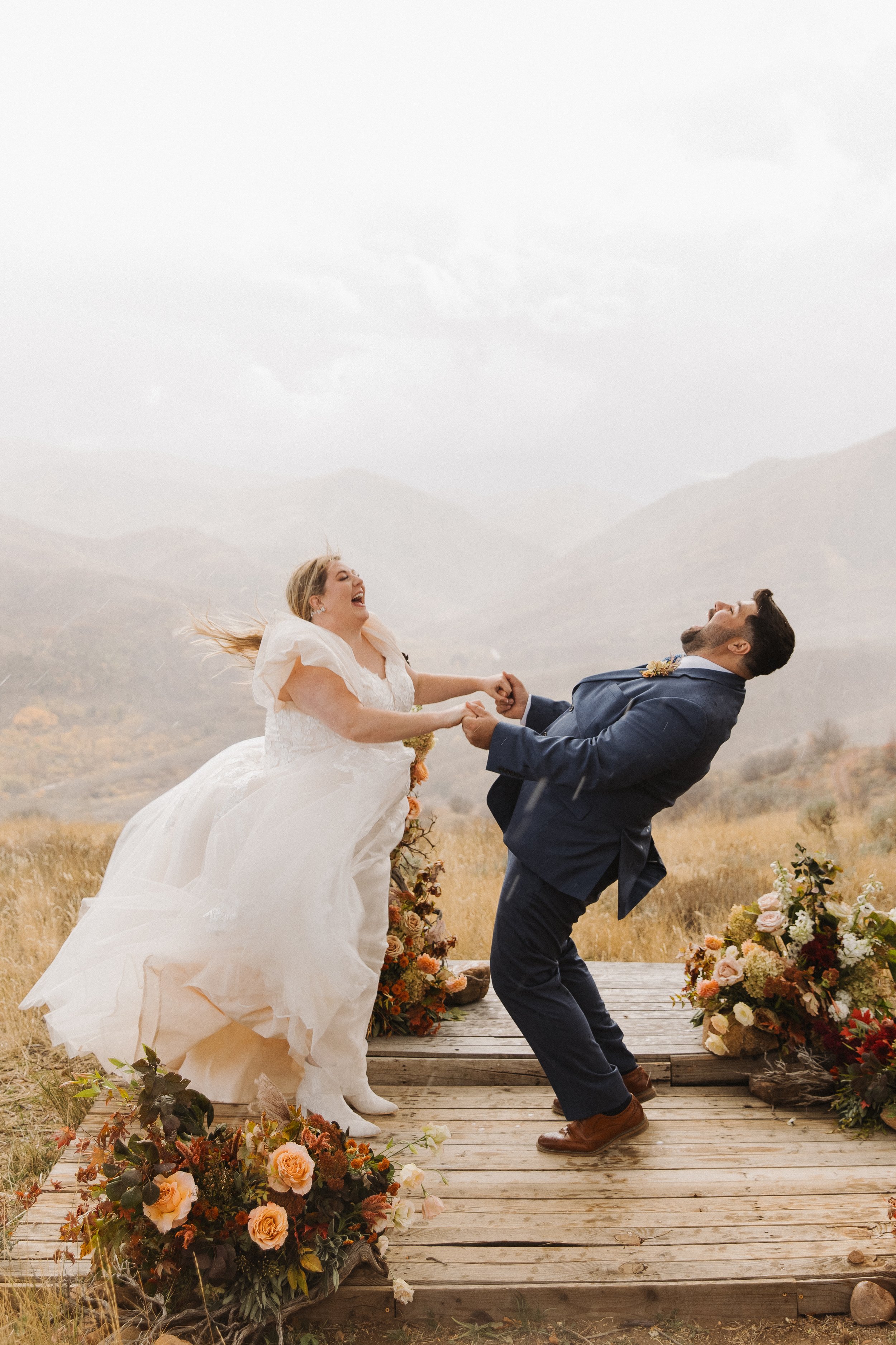 A joyful bride and groom laughing and dancing on an outdoor wooden platform surrounded by flowers, with a mountainous landscape in the background.