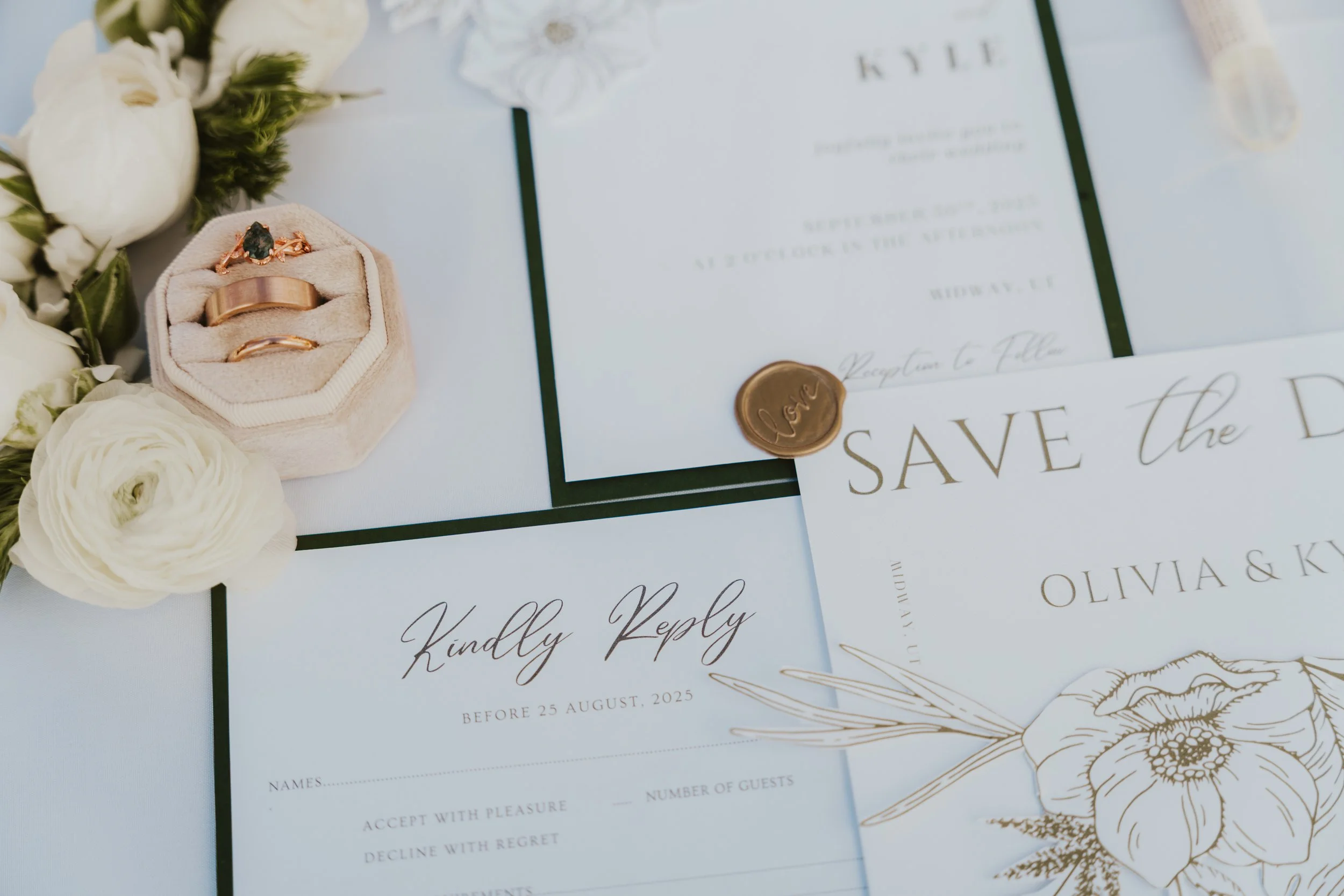 Wedding invitation cards with gold accents, a ring box with wedding rings and an engagement ring, white flowers, and a wax seal with the word "love" on it.