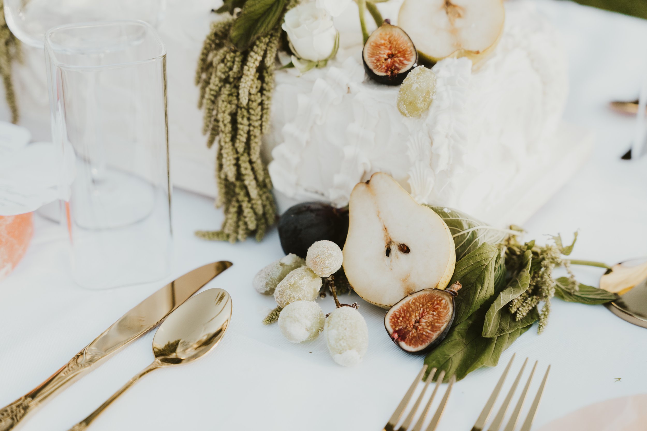 Decorative table setting with a white cake adorned with figs and greenery, gold cutlery, and a glass.