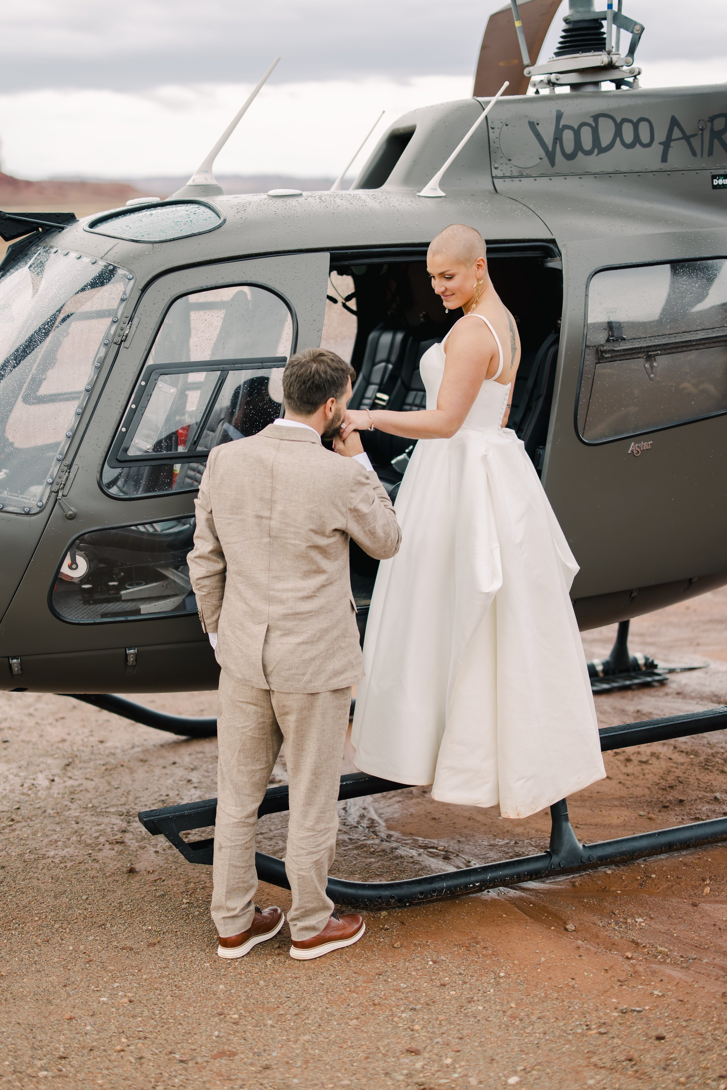 A couple dressed in wedding attire, the groom kneeling and holding hands of the bride, standing in the doorway of a helicopter in a desert landscape.