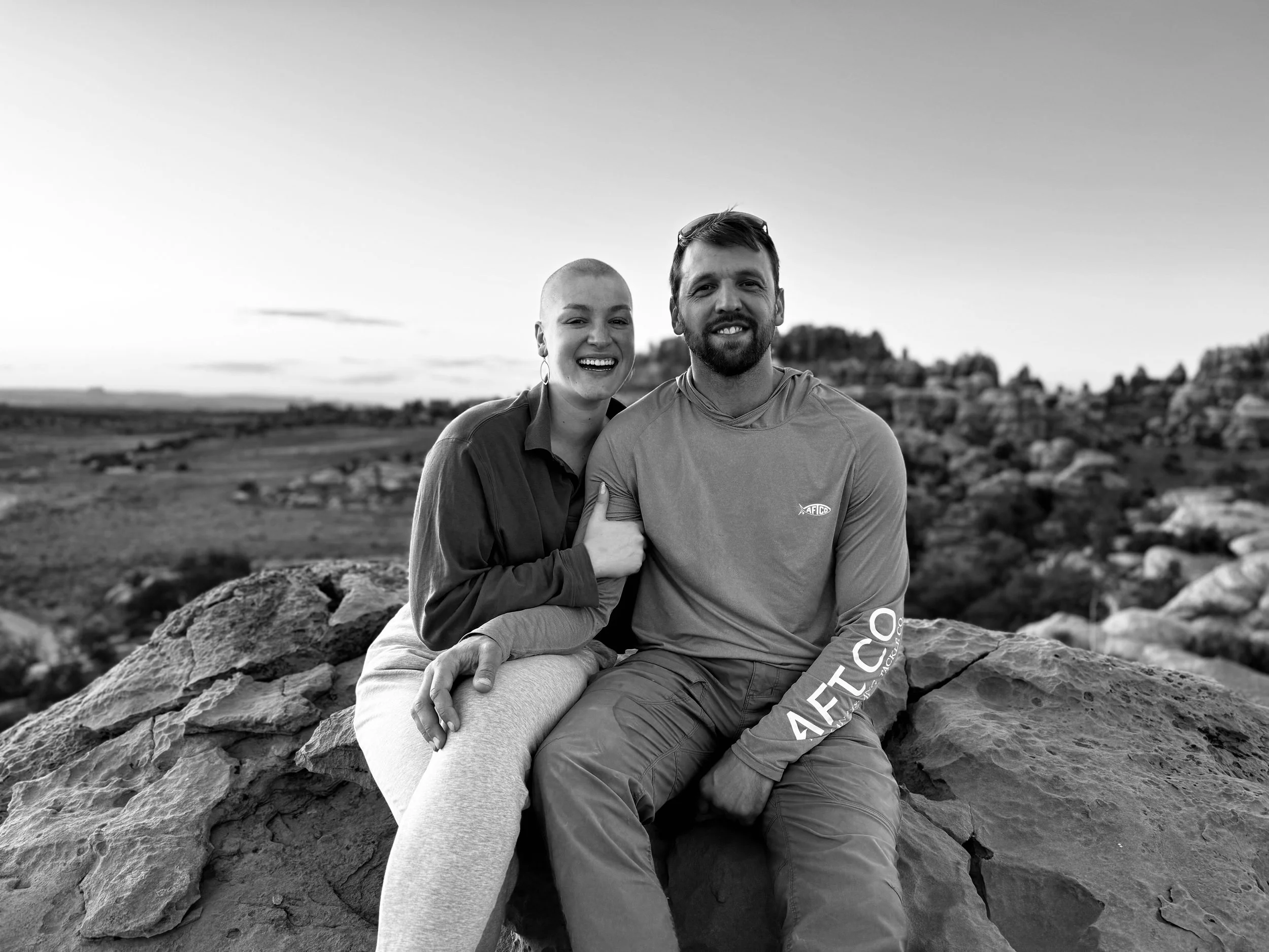 A smiling woman with a shaved head and a man with a beard sitting on a rock outdoors, with a landscape of rocks and a hill in the background, in black and white.