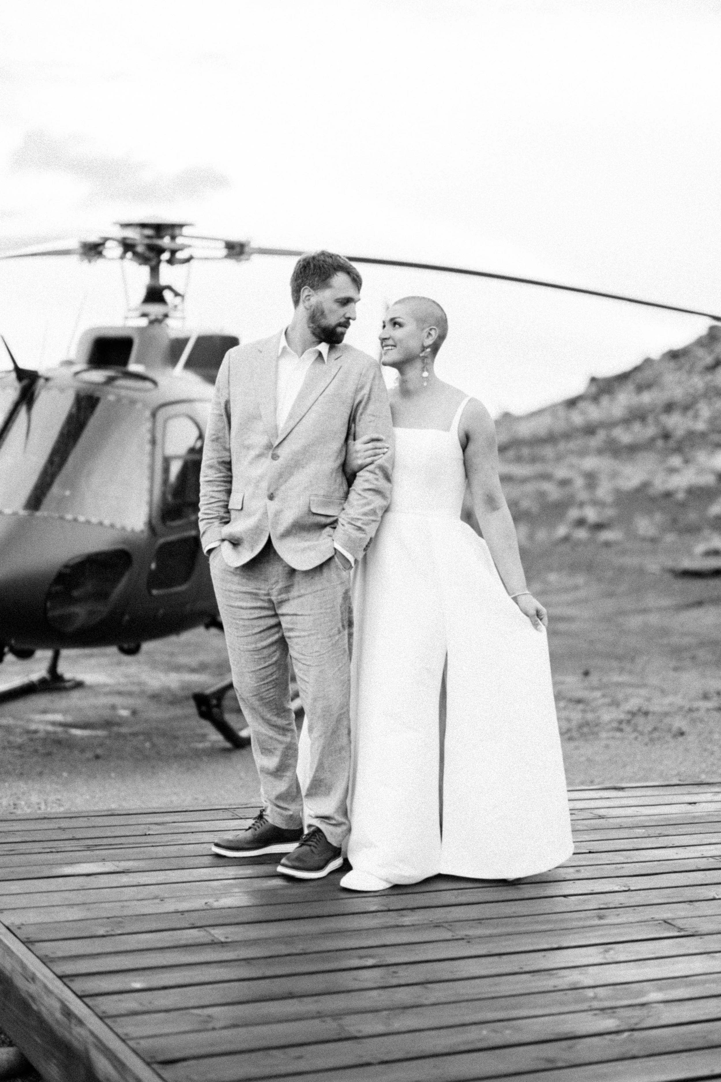 A couple dressed in wedding attire standing on a wooden platform with a helicopter in the background and a mountainous landscape.
