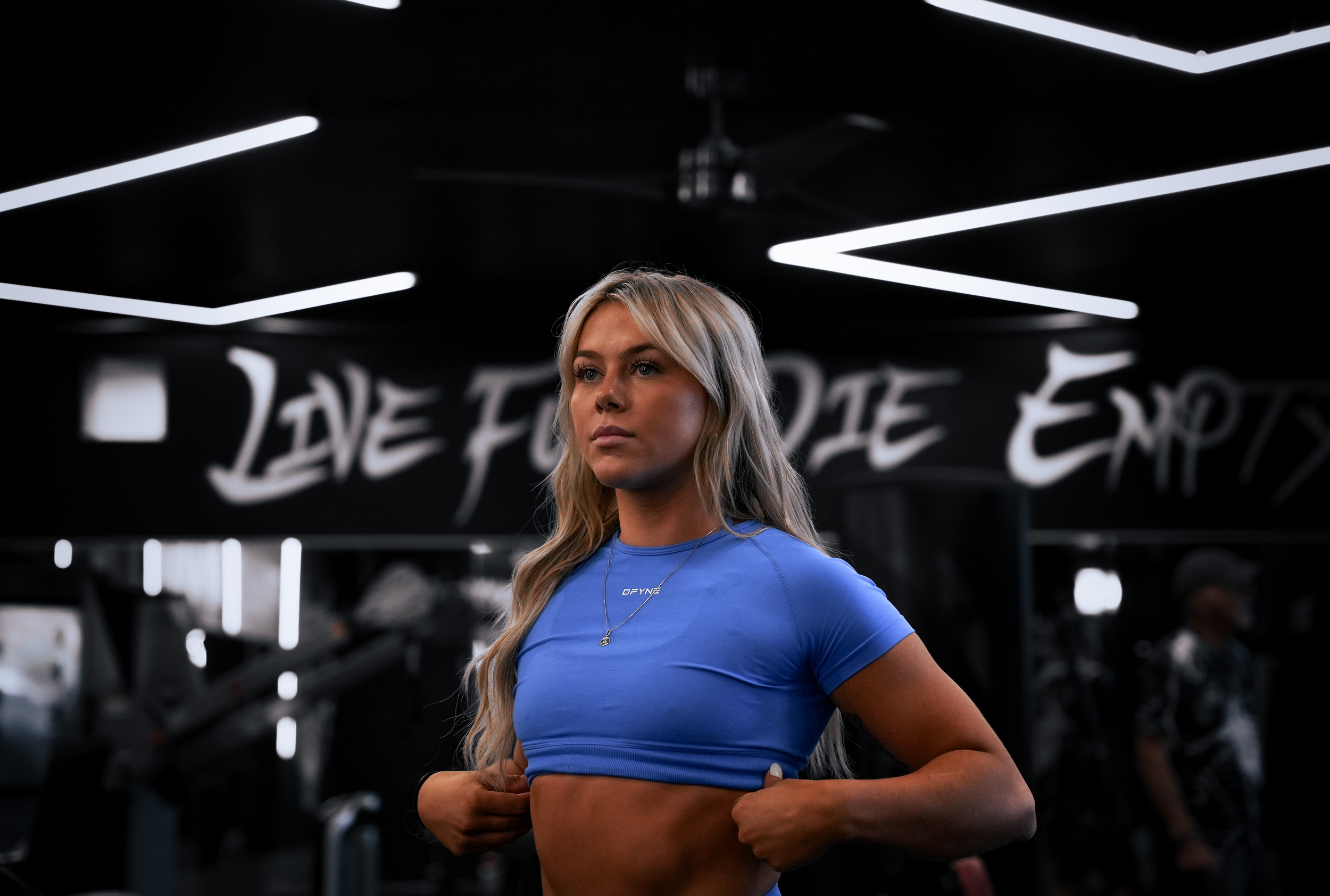 Woman in gym wearing blue sports top with motivational wall art in the background.