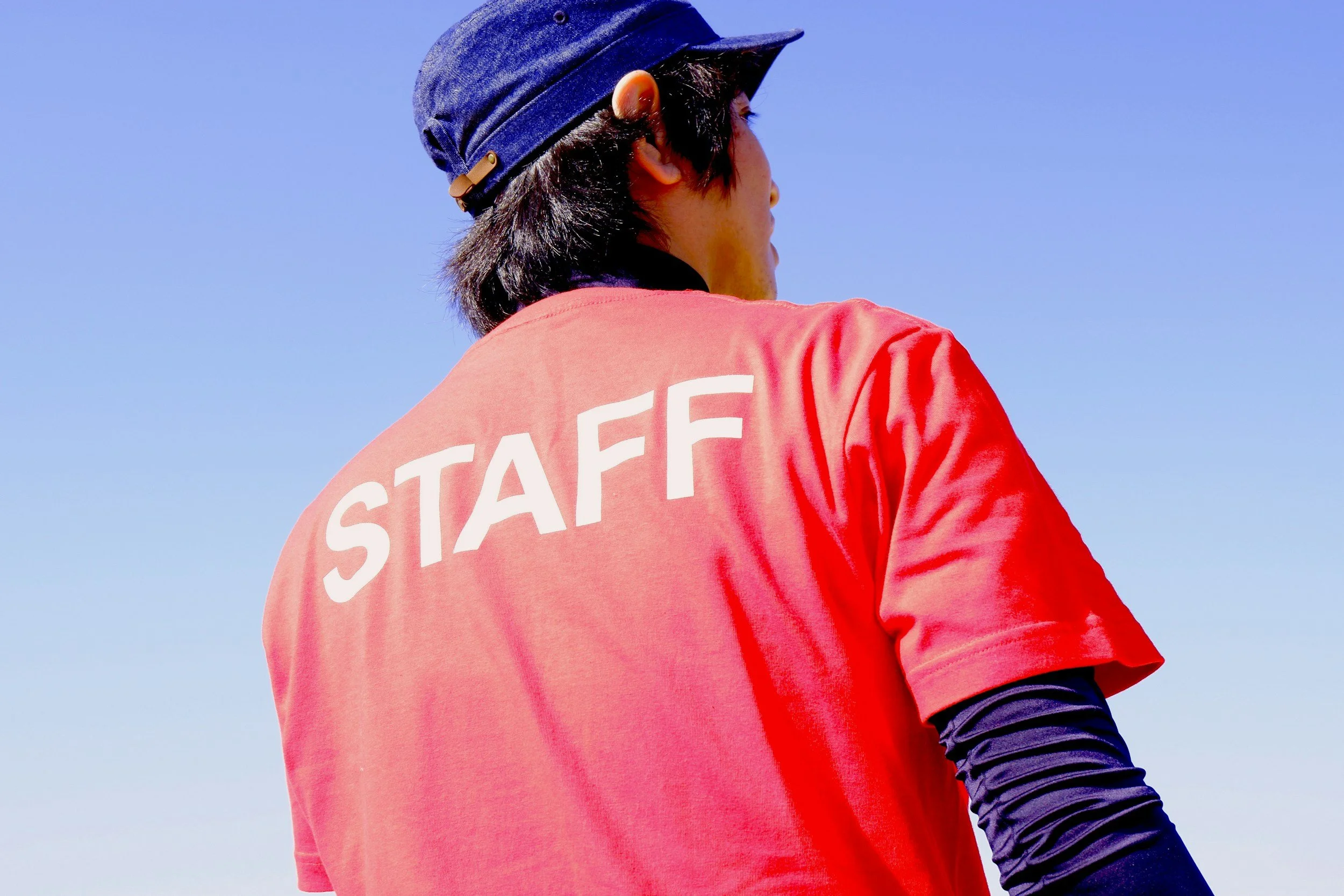 A person wearing a navy blue cap and a red shirt with the word "STAFF" on the back, standing outdoors against a clear blue sky.