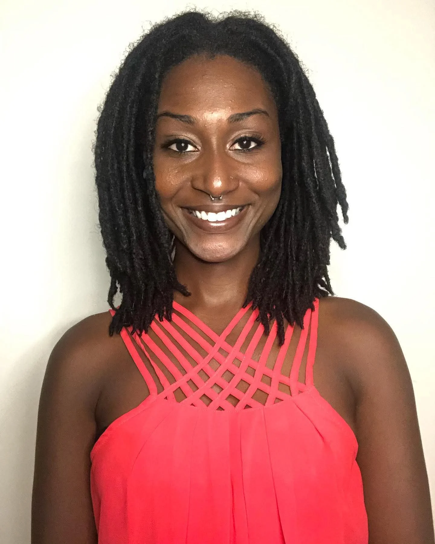 A woman with dark skin and shoulder-length dreadlocks wearing a red dress with a lattice design on the neckline, smiling in front of a plain white background.