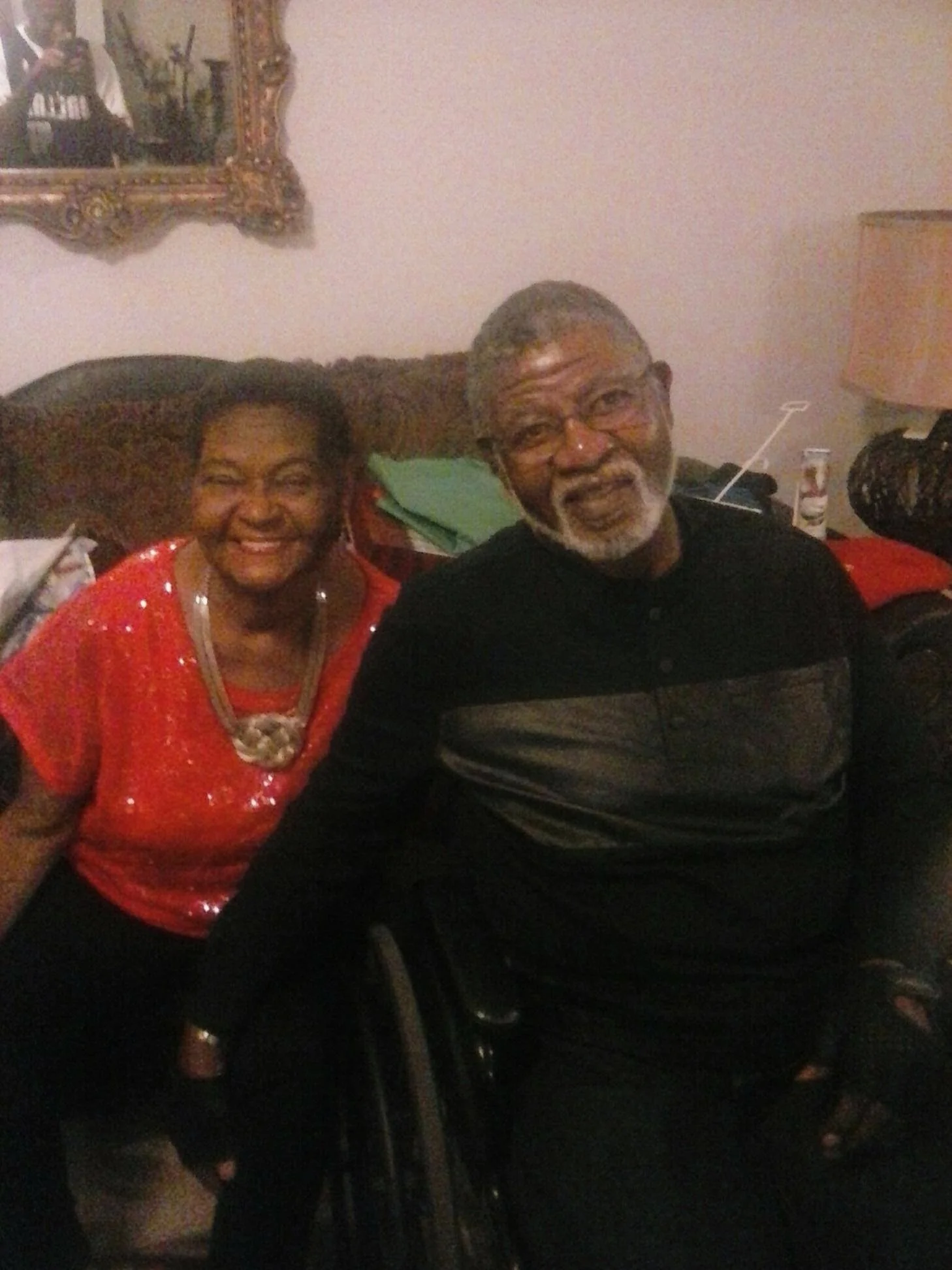 Smiling woman wearing a red sequined top and a man with glasses and beard sitting on a couch in a dimly lit room.