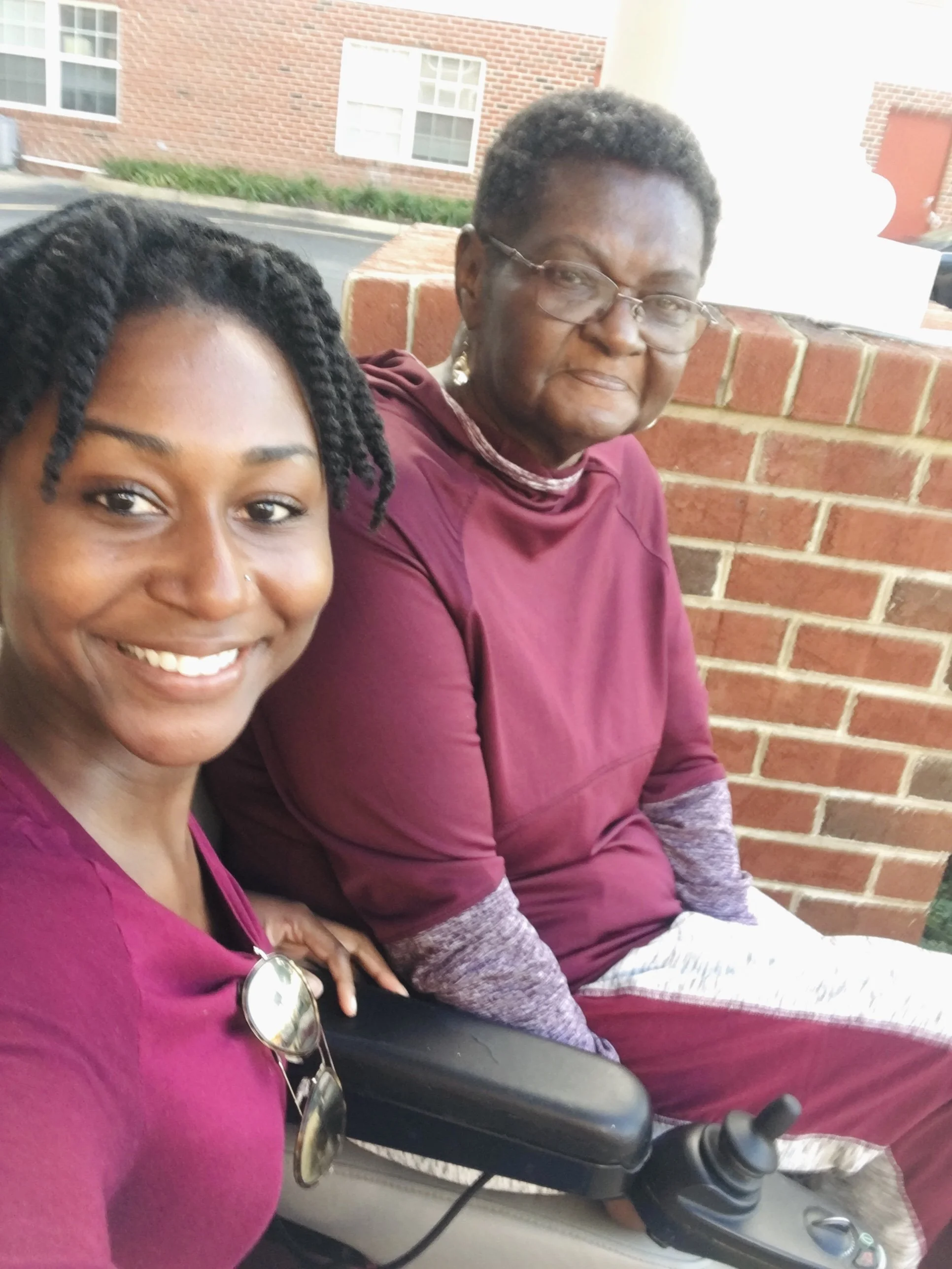 A woman with a nose piercing and glasses is smiling in a close-up selfie with an older woman in a maroon top and glasses, sitting against a brick wall outside.