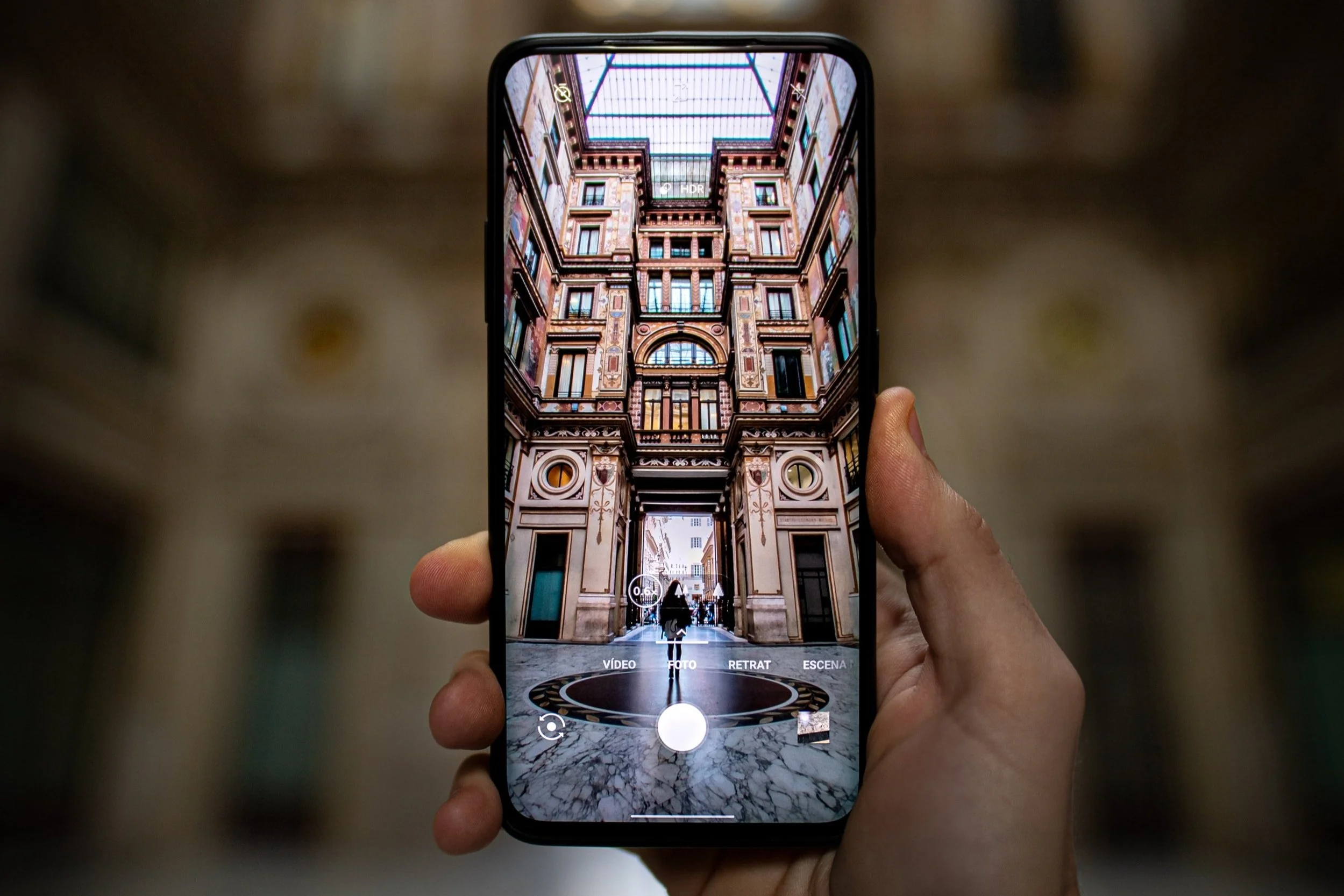 Person taking a photo of an ornate, multi-story building with a glass ceiling, using a smartphone camera app.