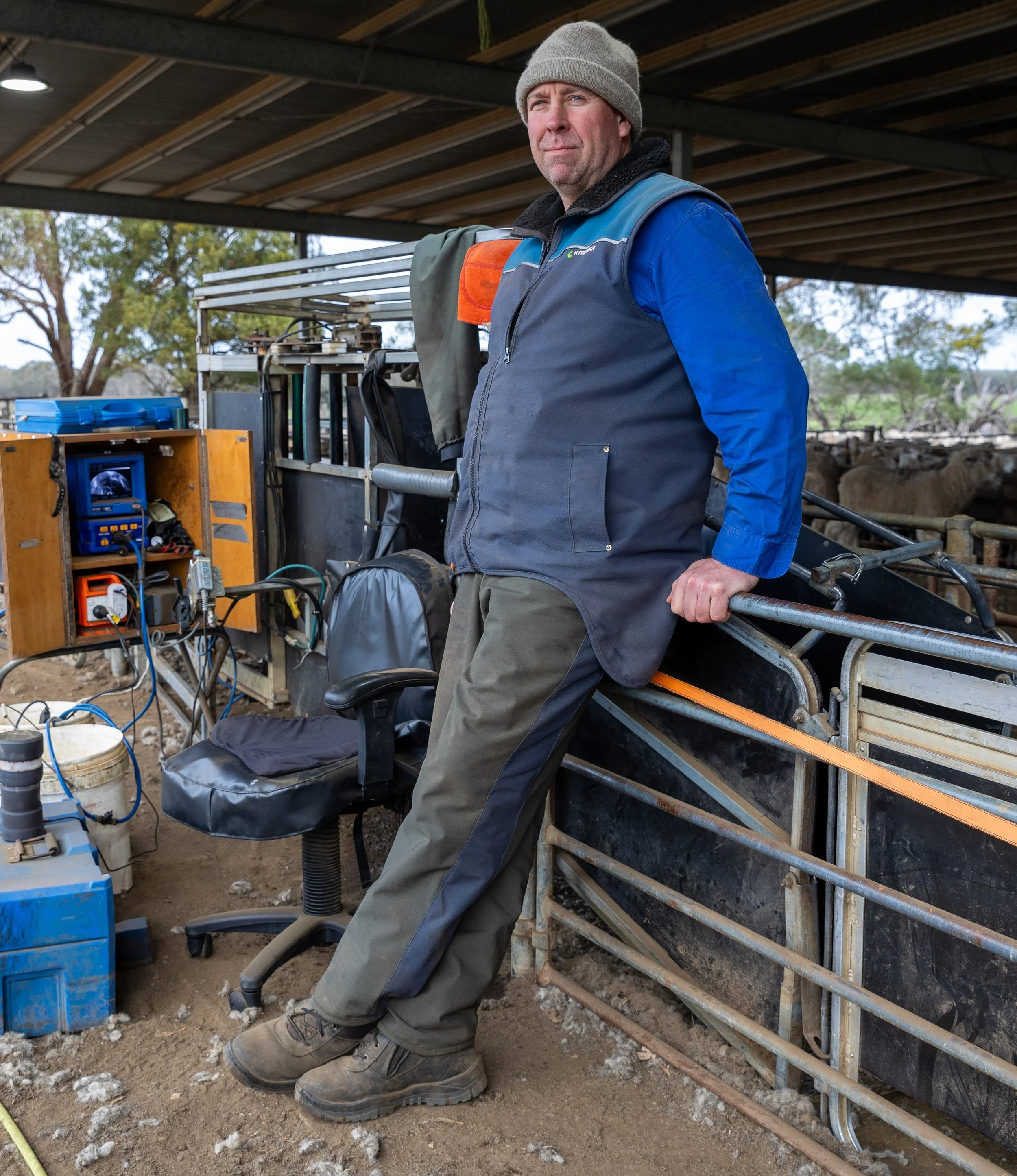 A man in work attire standing in a barn, leaning on a metal railing, with farming equipment and animals in the background.