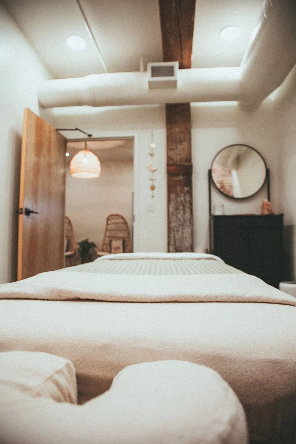 View of a cozy massage table in the foreground, a black dresser with a round mirror on top, a wooden door partially open revealing a sitting area with rattan chairs, a hanging woven lamp, and decorative moon and star hanging in the background.