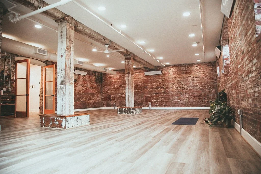 Spacious room with wooden floors, brick walls, ceiling lights, and large wooden columns, featuring a yoga mat and plants in the corner.